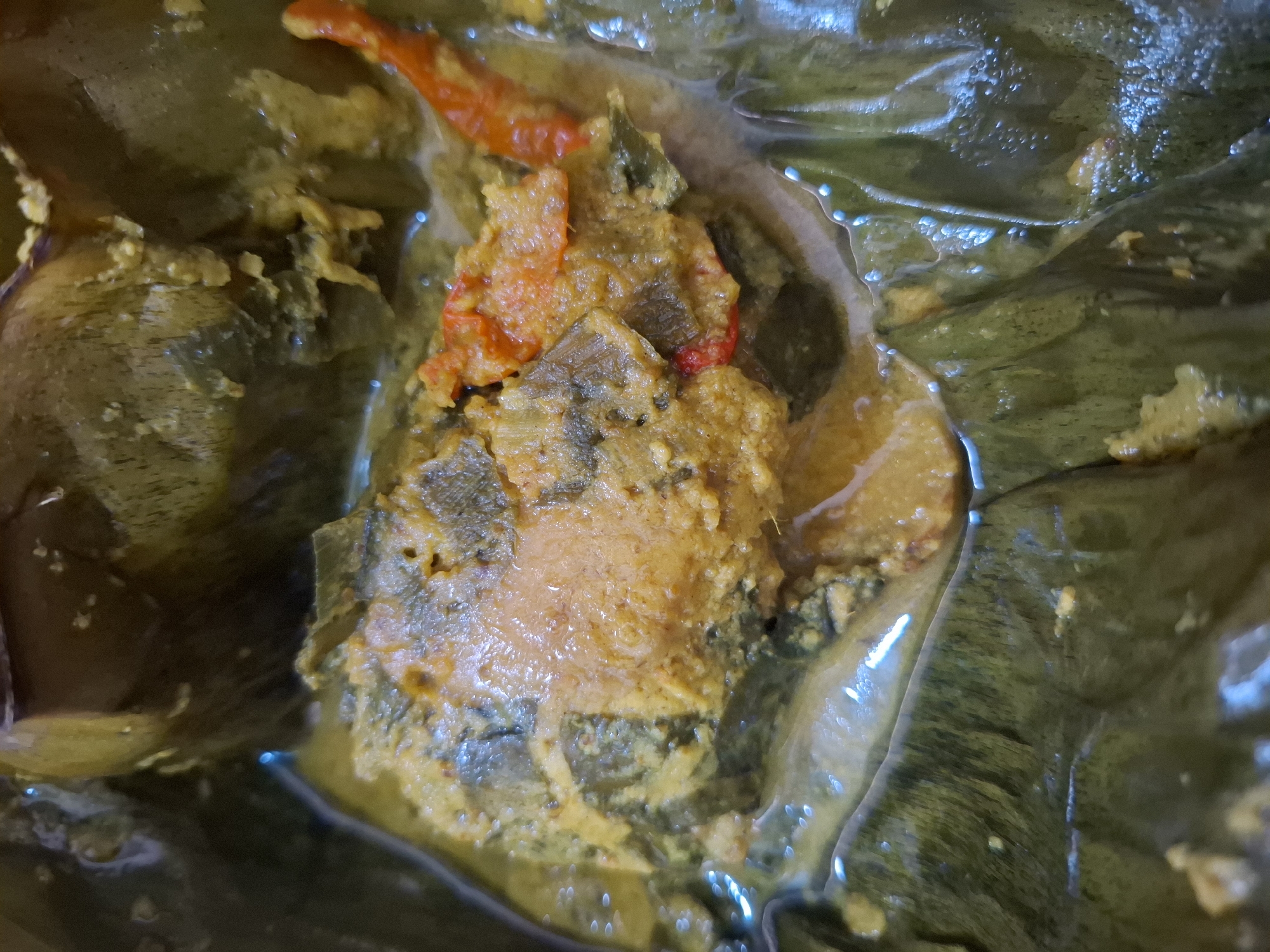The image is a close-up, top-down view of a dish prepared in banana leaves, commonly found in the city of Maleber, Indonesia. The dish appears to be a type of fish curry or steamed fish, with visible pieces of fish enveloped in a yellowish, creamy sauce. Red chili peppers are also present, adding a touch of color. The banana leaves, a traditional cooking and serving vessel, are clearly visible, providing a rustic and authentic presentation. The lighting suggests the dish is indoors, possibly in a kitchen or dining area. No people are visible in the frame.