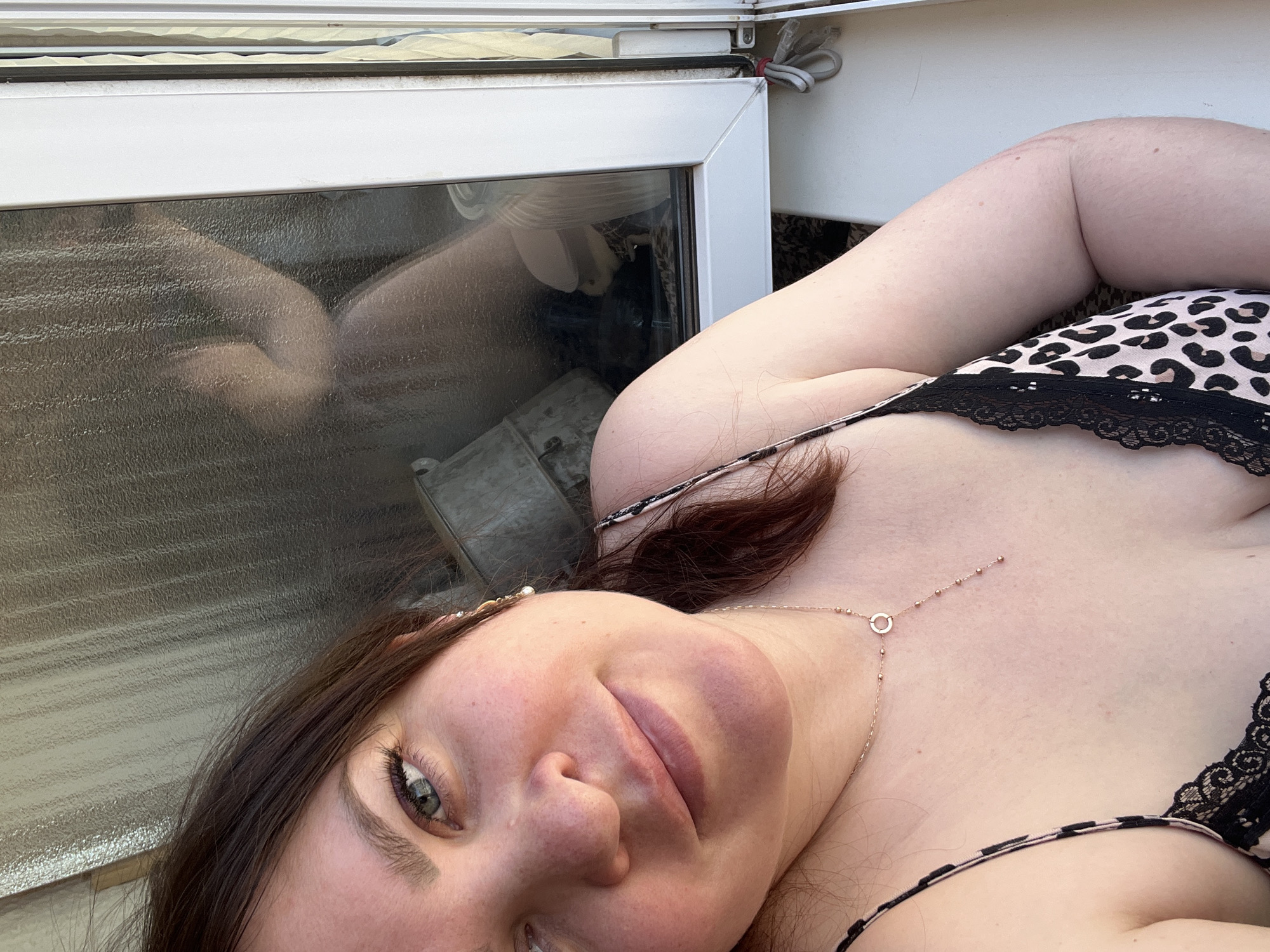 The image shows a woman with brown hair and fair skin wearing a leopard print top with black lace trim. She is lying down and looking at the camera with a slight smile. She is wearing a delicate gold necklace. To her left, there is a window with a reflection of what appears to be a bare leg, possibly hers, along with other indistinct objects. The background is indistinct, but it appears to be outside or near an exterior structure. There is no information to determine this is in the city of Noviy, Russia.