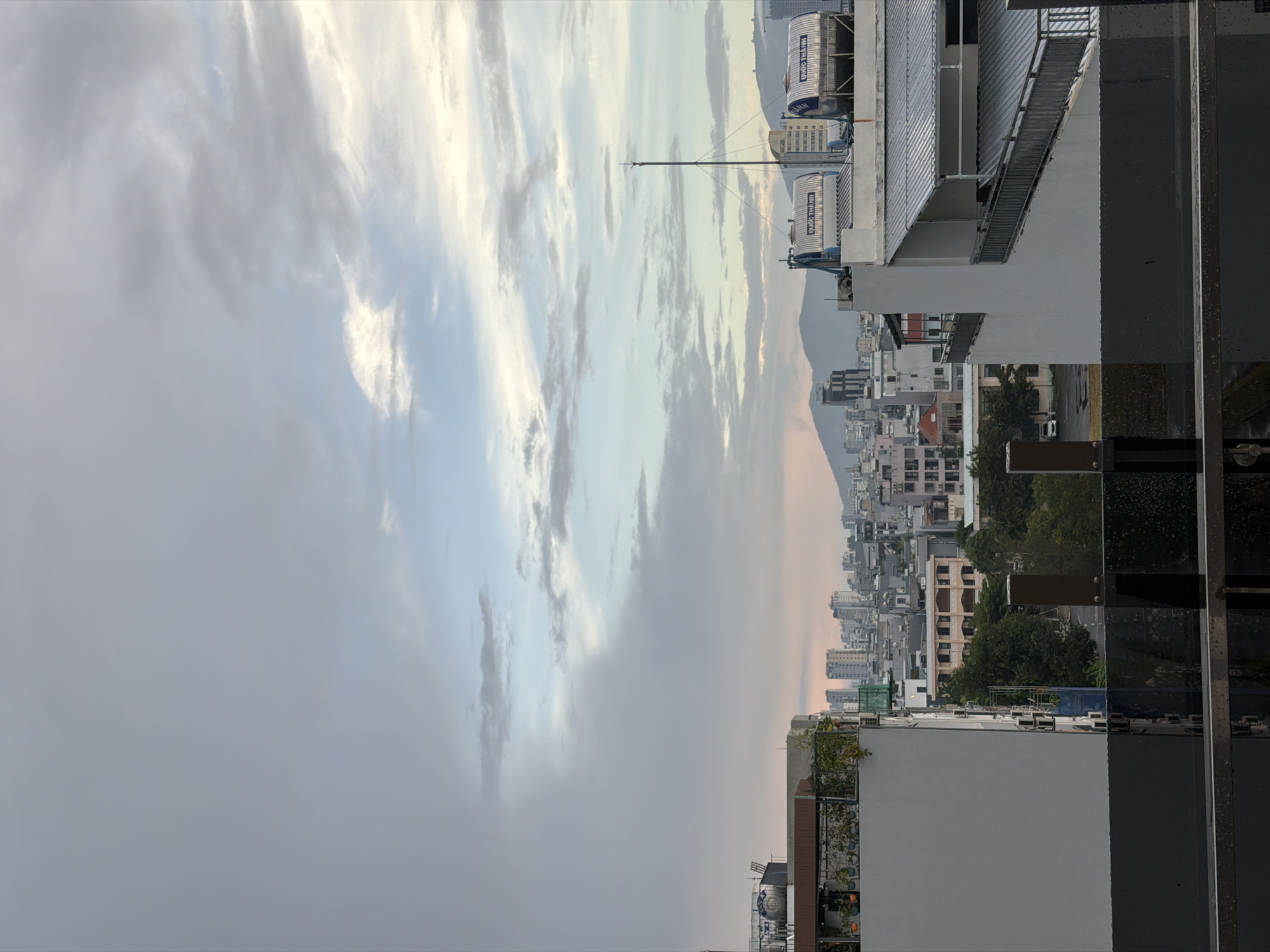 This image, taken in Đà Nẵng, Vietnam, depicts an urban landscape from a high vantage point, likely a rooftop. The scene is dominated by a vast expanse of sky filled with dramatic, streaky clouds that transition from a pale, overcast gray on the left to hues of white, blue, and soft pinks on the right, suggesting either early morning or late afternoon.
Below the sky, the cityscape unfolds. Buildings of varying heights and architectural styles are visible, densely packed together. Many of them appear to be residential or commercial blocks, with light-colored facades and numerous windows. Some rooftops are adorned with greenery, indicating urban gardens or terraces. In the middle ground, large cylindrical water tanks are prominently situated on a building's roof, emblazoned with Vietnamese text. One tank clearly reads "QUỐC THÀNH" and "SON". Another smaller tank, also with "QUỐC THÀNH" is visible below it. Further into the distance, a hazy mountain range forms the backdrop to the urban sprawl.
The foreground is occupied by the edge of a modern building with dark, reflective surfaces, possibly glass, and some exposed metal structural elements, suggesting a contemporary construction. The overall impression is one of a bustling city under a dynamic sky, with a sense of distance and scale. The lighting and cloud formations suggest a period of transition in the day, possibly near sunset or sunrise, creating a slightly subdued or ethereal atmosphere.