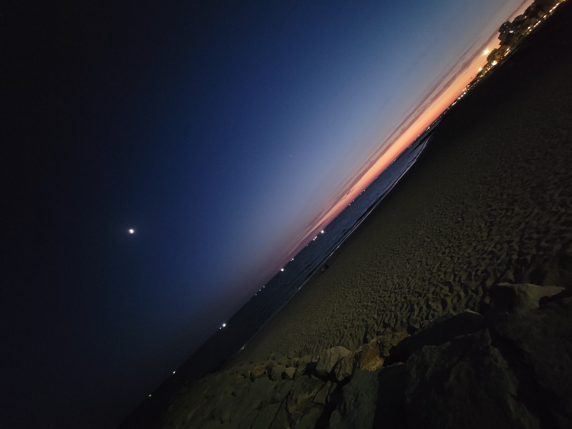 The image depicts a night scene on a beach in Quarteira, Portugal. The perspective is from ground level, looking out towards the sea and sky. A rocky embankment is in the foreground on the left, with dark shadows cast on the sand. The beach itself is sandy and appears mostly empty.
The sea is visible to the right of the embankment, with gentle waves. Along the horizon, there is a faint band of light, suggesting the last remnants of twilight or artificial lights from a distant town. Several small, bright lights are scattered along the waterline, which could be distant boats or lights from buildings.
The sky is a deep gradient of blues, transitioning from a dark, almost black blue at the top to a lighter, more muted blue near the horizon. A crescent moon is visible in the upper right portion of the sky, along with a few faint stars. The overall impression is one of a peaceful, late evening on the coast. The angle of the shot is tilted, giving a dynamic feel to the scene.