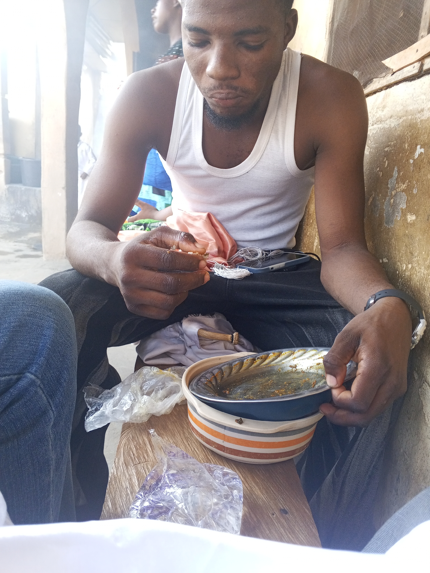 A medium shot reveals a man with dark skin, clad in a white tank top and dark pants, seated outdoors. The setting is presumably Lokoja, Nigeria, though specific landmarks are not visible. The man appears to be eating from a bowl with orange and white stripes, which sits on a wooden surface. His left hand holds a piece of food, while his right hand steadies the bowl. Various plastic bags and cloths are scattered around the bowl on the wooden surface. A portion of a person wearing blue jeans is visible on the left side of the image. The background shows a building with a light-colored wall. The man has a black beard and is wearing a watch on his right wrist. A mobile phone is lying on his lap.