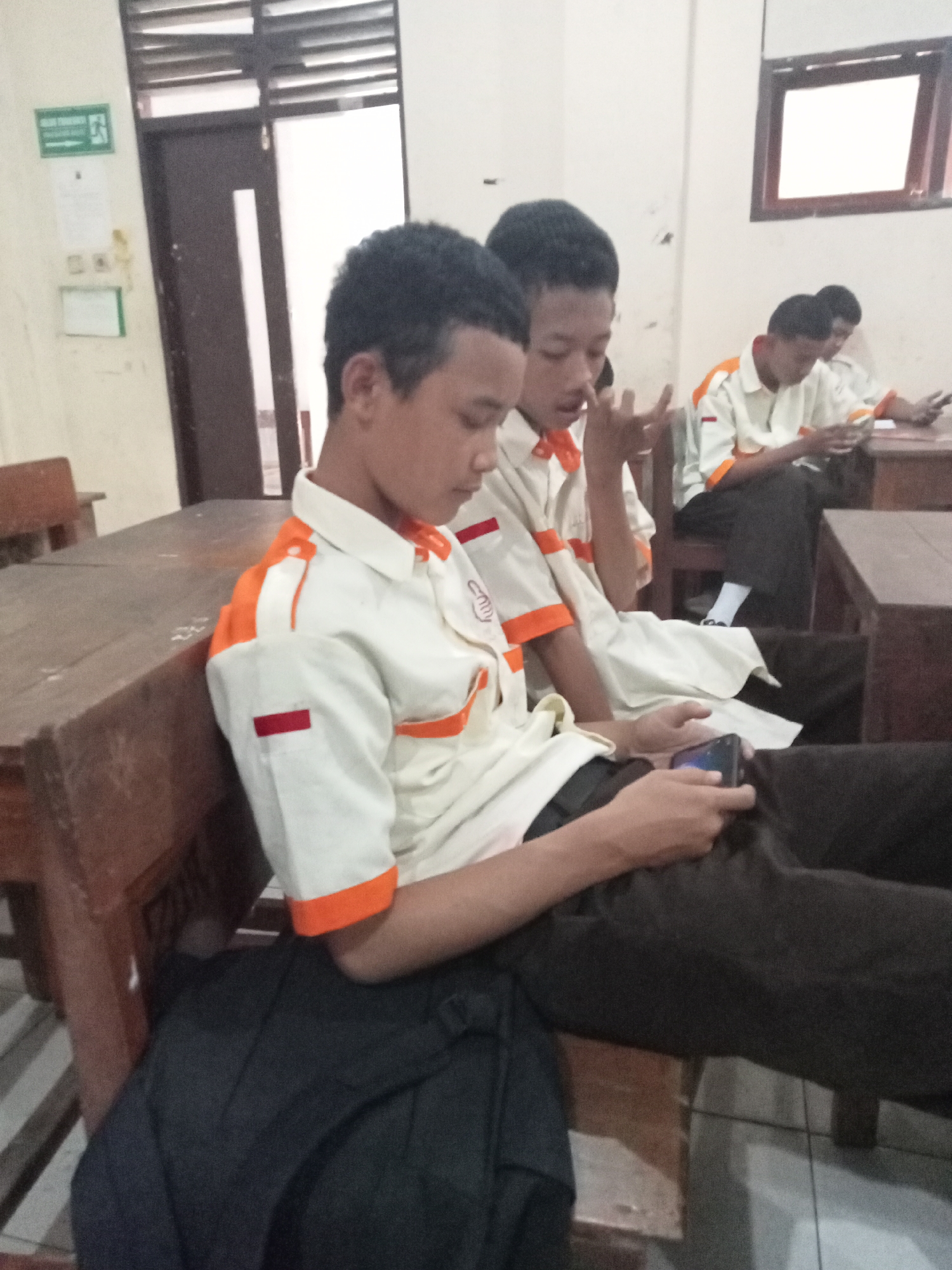 The image depicts an indoor scene with several young individuals wearing school uniforms, suggesting a classroom or school common area. The uniforms consist of light-colored shirts with orange accents and dark trousers. The individuals appear to be of Indonesian descent. One person in the foreground is holding a mobile phone and looking at it. Another person next to them is also looking at a phone. In the background, two more individuals are seated, one of whom is also engaged with a phone. The environment includes wooden desks and chairs, a doorway with a glass panel, and a window with blinds. A green safety sign with an exit symbol is visible on the wall. The overall impression is one of a relaxed atmosphere within a school setting, where students are using their mobile devices.