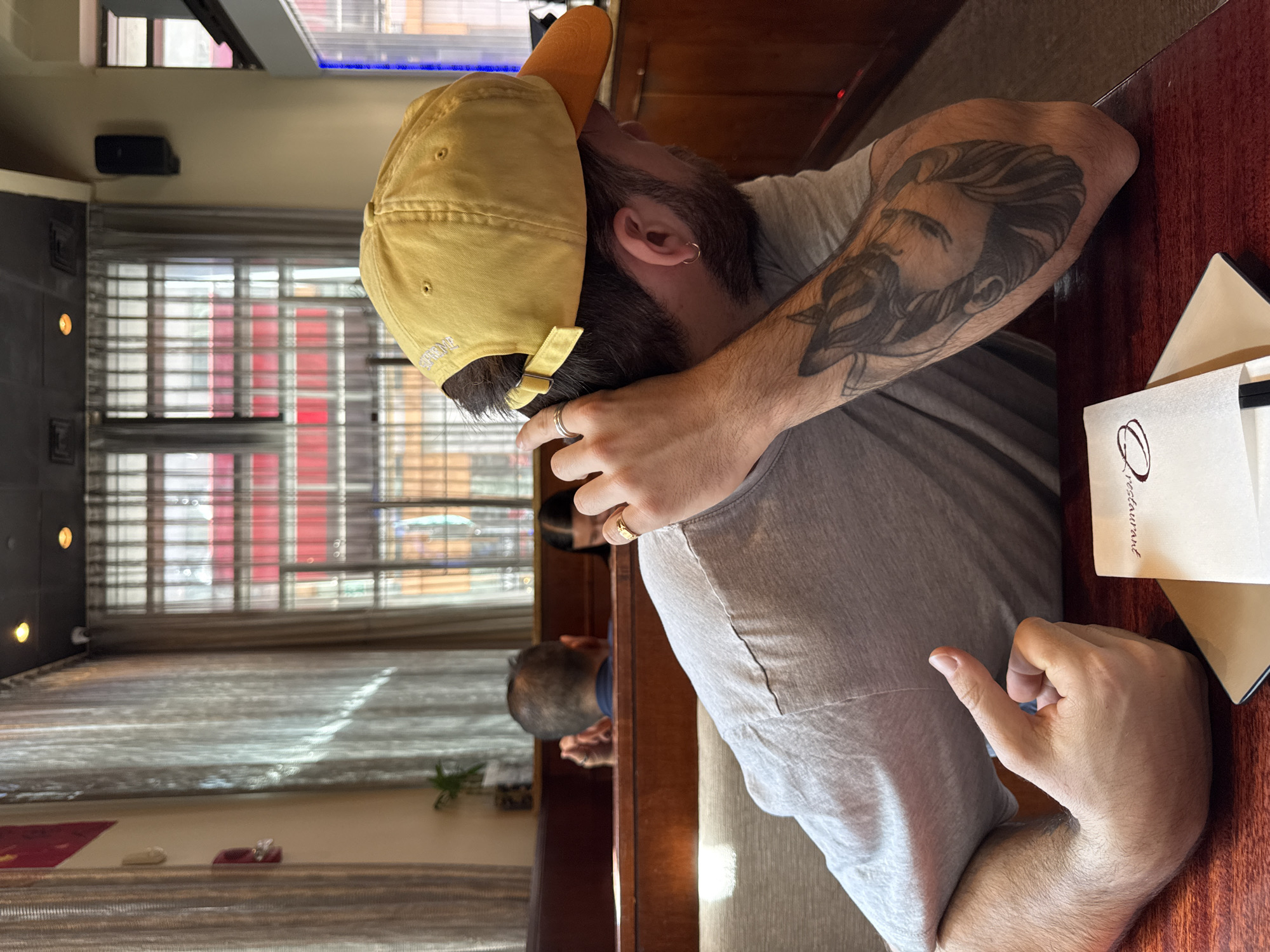 A man wearing a yellow baseball cap and grey shirt is sitting at a table in a restaurant. He has a tattoo on his right arm. The restaurant appears to be located in Boston, as evidenced by the "restaurant" wording on a white paper wrapper on the table. Outside the window, there are buildings with red and white exterior designs. Another person can be seen in the background.