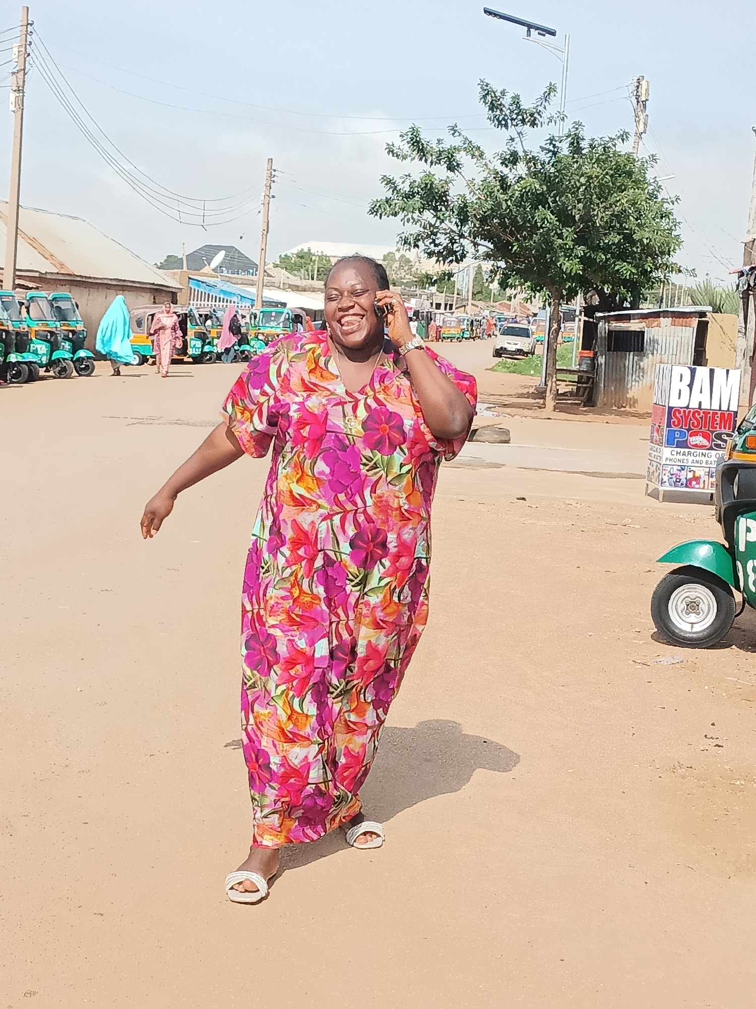 In an outdoor scene on a sandy road in Jos, Nigeria, a Black woman wearing a vibrant, floral-print dress and white sandals is captured mid-stride while talking on her cell phone. Her expression is one of joyful laughter, with her mouth open and eyes crinkled. Her left arm is extended slightly to her side, and her right hand holds the phone to her ear.
The background depicts a busy street scene. A row of teal-colored auto rickshaws, commonly known as "keke napeps," are parked along the left side of the road, suggesting a transportation hub. Several people are visible near the rickshaws, some dressed in traditional attire including a woman in a blue hijab. Further down the road, other vehicles, including a silver car, are present.
On the right side of the image, a small, corrugated metal structure stands, with a sign above it that reads "BAM SYSTEM POS." Next to this structure, the front wheel and fender of a green auto rickshaw are visible. Power lines and utility poles crisscross the sky, indicating an urban environment. A lone tree with green foliage stands near the center of the frame. The overall impression is a snapshot of everyday life in a bustling part of Jos.