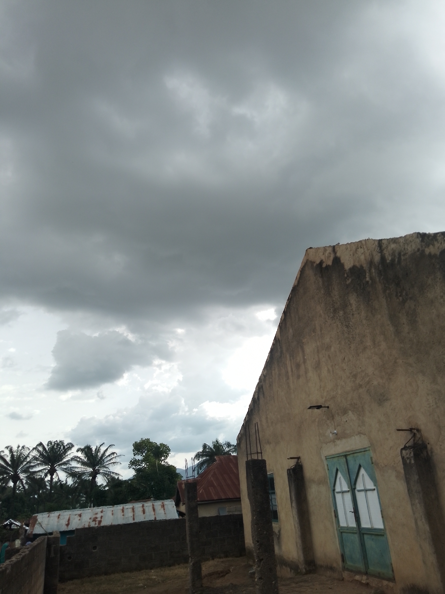The image depicts a cloudy sky over the town of Serti, Nigeria. In the foreground, a light-colored building with a rough texture and dark water stains on its facade dominates the right side of the frame. This building has a steeply pitched roofline, and a double-door painted teal is visible on its side. To the left of this building, a wall constructed of dark bricks is present, and beyond that, several structures with corrugated metal roofs can be seen. In the background, lush green trees and palm trees are scattered throughout the landscape, suggesting a tropical or sub-tropical environment. The overall atmosphere is subdued due to the overcast conditions, with the sky filled with heavy, grey clouds.