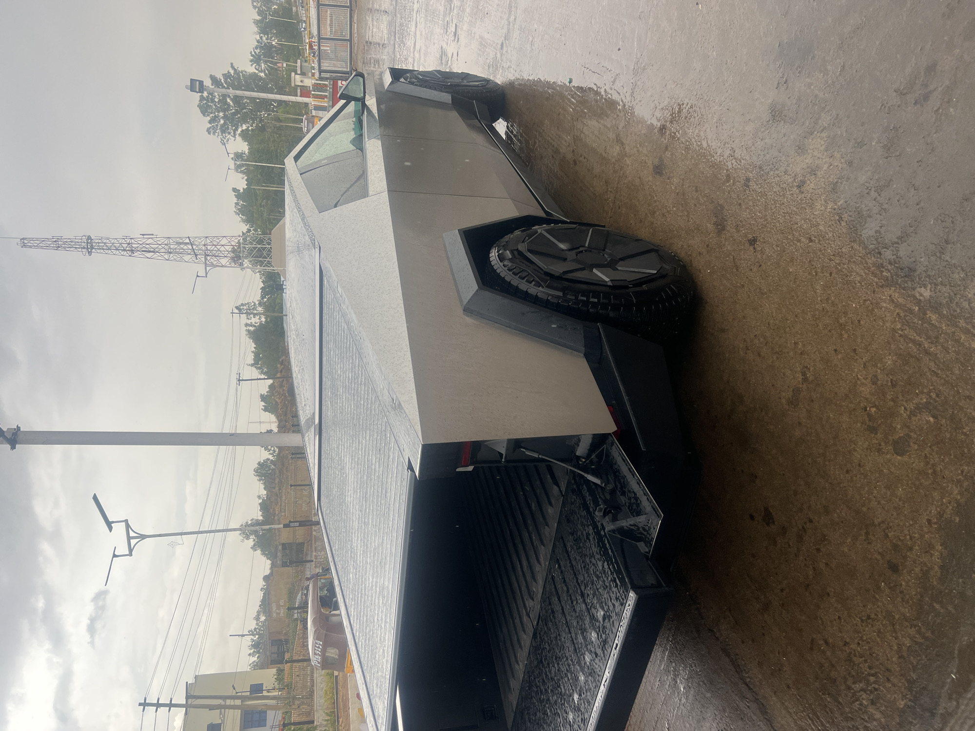 The image shows a Tesla Cybertruck parked on a wet surface in an outdoor setting. The truck is silver and has a distinctive geometric design. The ground is damp, reflecting the overcast sky. A tall metal tower is visible in the background, along with trees, utility poles, and buildings. A vehicle can also be seen in the background. The image appears to be taken in Jos, Nigeria.