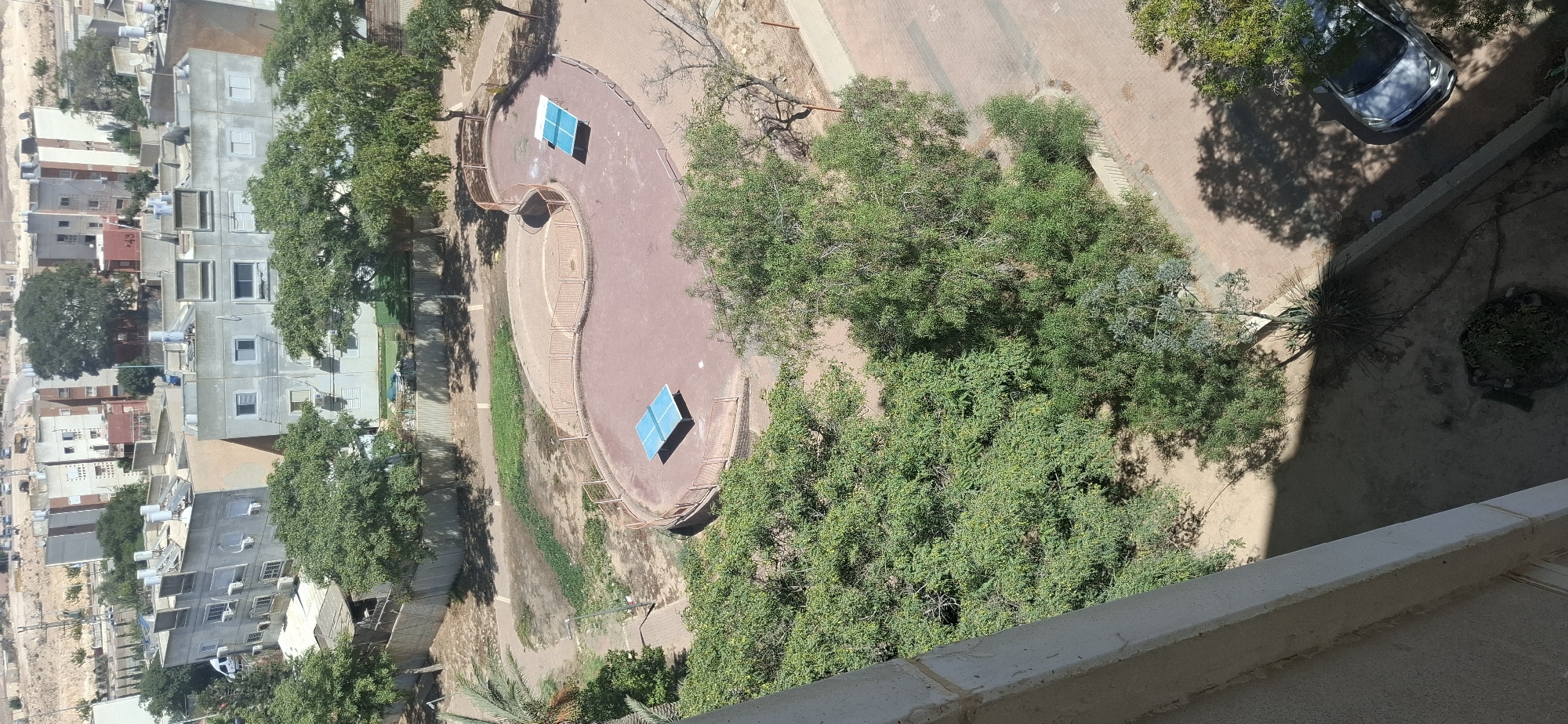The image captures an aerial view of a public park area in Dimona, Israel. In the center of the frame, a kidney-shaped, paved area with a red surface is visible. Two blue ping pong tables are positioned on this surface, one closer to the foreground and the other further back. The area is enclosed by a low, orange metal fence. Lush green trees surround the paved area, with some casting shadows on the ground. Beyond the park, several multi-story apartment buildings with light-colored facades and solar water heaters on their roofs are visible, characteristic of urban residential areas in Israel. In the lower right portion of the image, a silver car is parked on a paved walkway adjacent to the park. The surrounding environment appears dry and arid, consistent with the climate of Dimona.