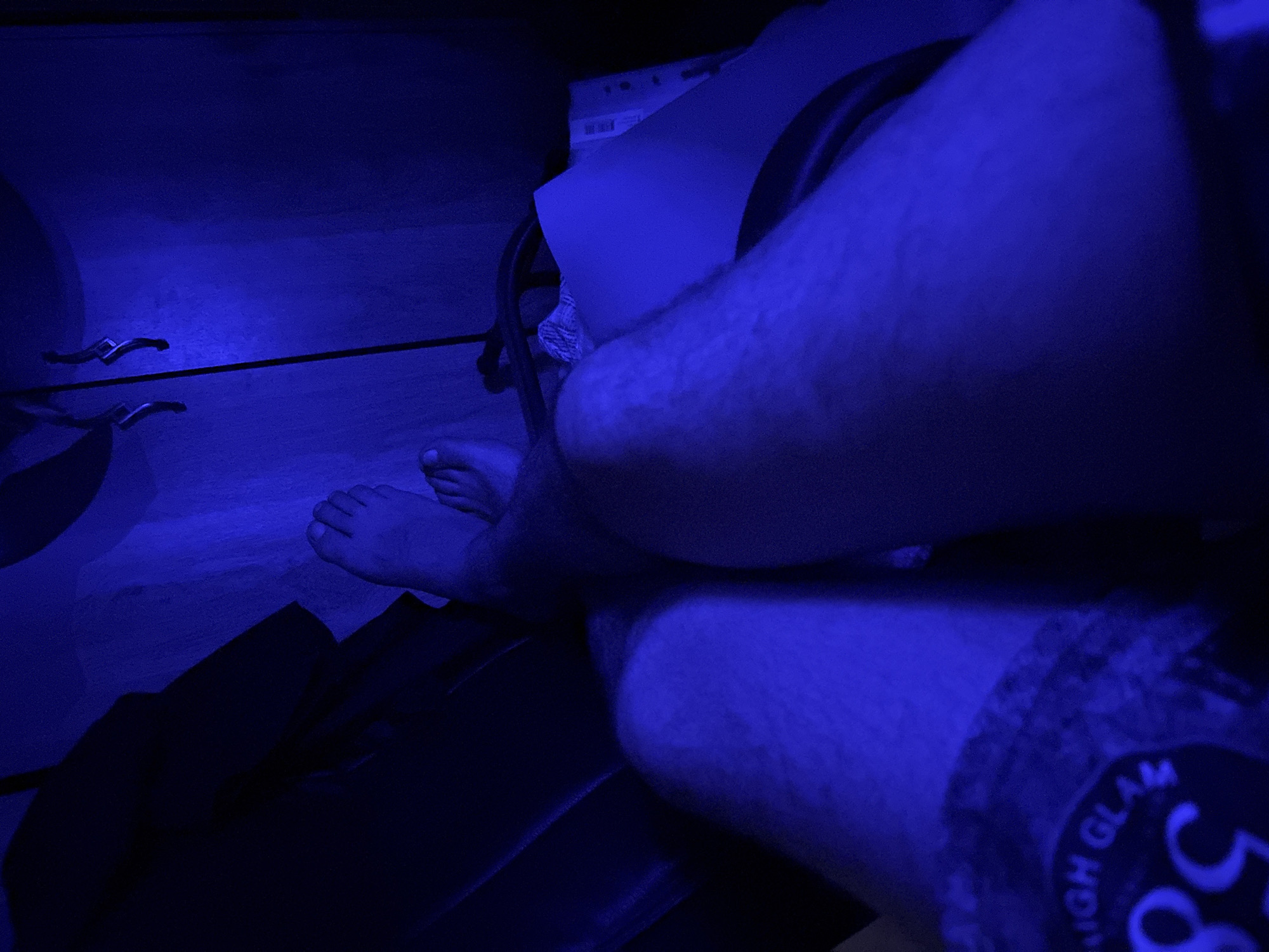 The image captures a close-up view of a person's legs and feet, bathed in a strong blue light, creating a dark and atmospheric setting. The person appears to be sitting, with their legs bent at the knees. One foot is clearly visible, bare and resting on what seems to be a dark, textured surface. The other leg is mostly obscured, but hints of clothing or fabric are present.
In the background, the blue light illuminates a horizontal surface, possibly a desk or shelf, with two metallic objects resembling levers or handles positioned on it. Further back, a section of a white sheet of paper or a thin board is visible, also under the blue light. The overall scene suggests an indoor environment, perhaps a room or a confined space, where the blue light is the dominant source of illumination. The shadows are deep, and the blue hue lends a somewhat mysterious or futuristic feel to the image.
There is some visible text on the lower right portion of the image, appearing on what seems to be fabric. The text is partially obscured and difficult to read clearly due to the lighting and angle, but "HIGH GLAM" and the number "58" are discernable.
The activity taking place is not explicitly clear, but the posture suggests the person is engaged in something while seated. There are no clear indicators of time of day or weather conditions. The location context provided is "Qarağandı city administration, Kazakhstan," which is a factual detail about the setting. The emotional tone is difficult to ascertain definitively due to the lighting and close-up nature of the shot, but the blue light might evoke a sense of calmness, introspection, or even a slight tension depending on interpretation.