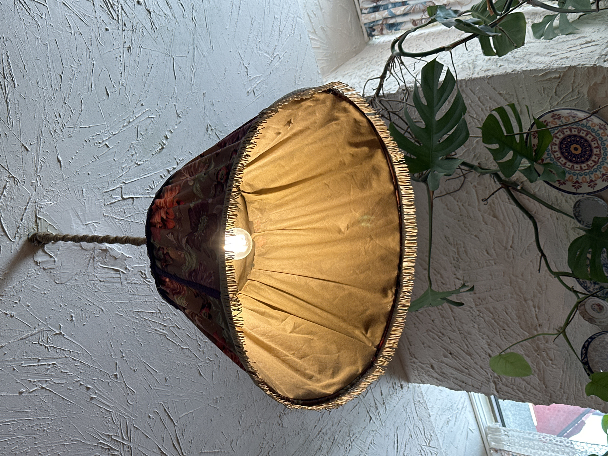 A lampshade with an illuminated bulb hangs from a textured wall in what appears to be an interior space. The lampshade has a patterned outer layer and a tan-colored lining with a fringed edge. A rope-like cord connects it to the wall. A monstera plant is visible in the background, along with decorative plates on the wall. A window with a lace curtain is partially visible below the wall. The scene suggests a cozy, possibly bohemian, aesthetic.
The location is Tbilisi, Georgia.
