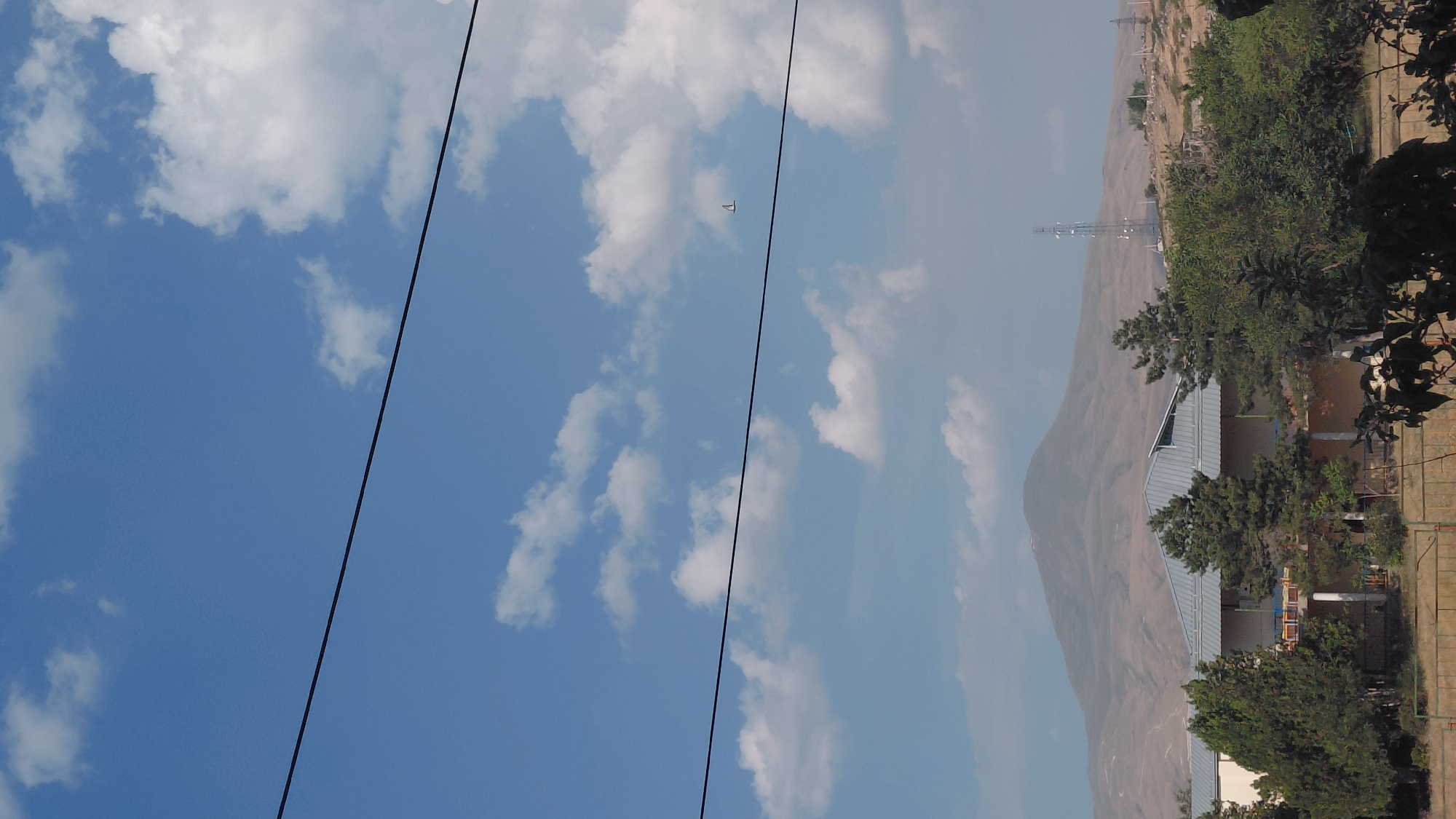 The image depicts a wide view of a landscape with a clear blue sky dotted with white clouds. In the foreground, the roofline of a building with a metallic roof is visible, bordered by various trees. In the mid-ground, a range of hills stretches across the horizon, culminating in a large, rounded mountain. A cell tower is also present on the right side of the frame. In the distance, the sky features two thin, dark horizontal lines, likely power lines, and a small, delta-shaped object, possibly a kite or a distant aircraft, can be seen in the upper right quadrant. The scene appears to be set in a semi-rural or suburban area.