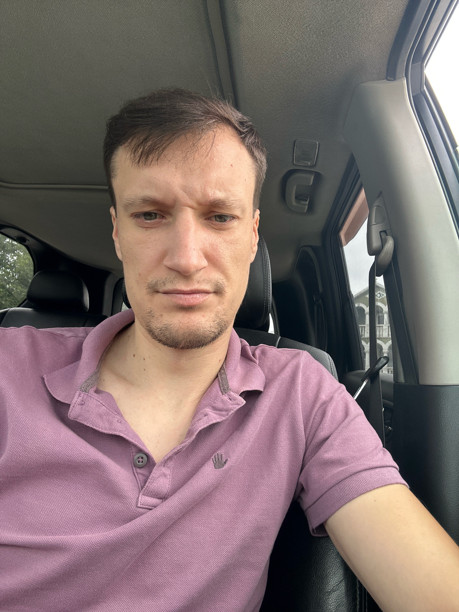 A man is sitting in the driver's seat of a car, facing forward and looking at the camera. He has brown hair, a light complexion, and is wearing a pink polo shirt. His expression is neutral. The interior of the car is visible, including the dashboard, steering wheel, and a portion of the passenger seat. The image appears to have been taken outdoors during the day, as evidenced by the natural lighting and the view of trees and buildings in the background. It is not possible to determine the location within Shatava, Ukraine from the provided image.