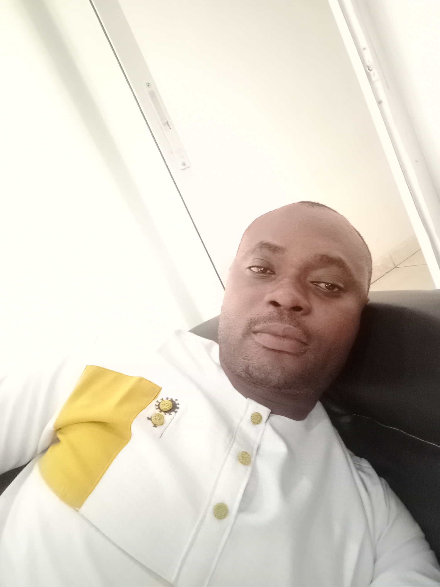A man is in Gwarinpa, Nigeria. The man has dark skin and is wearing a white shirt with gold buttons and a yellow pocket square. The shirt has an insignia. He is sitting in a black leather chair. The background is a white wall with a window.