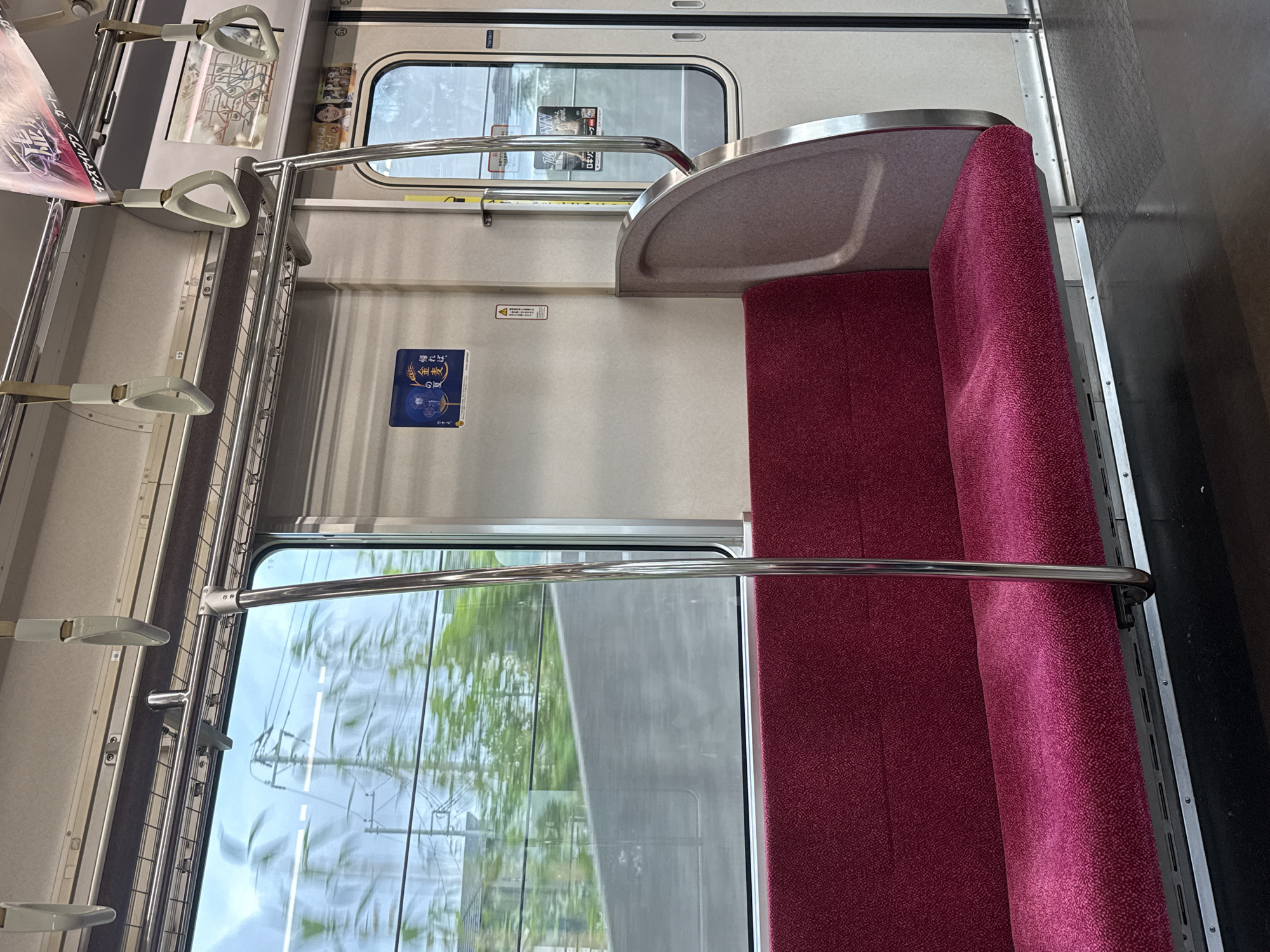 The image shows the interior of a train car. The seats are upholstered in a textured magenta fabric, and there is a silver metal pole running vertically in front of the seats. The window on the left shows a blurred view of green trees and utility poles, indicating the train is in motion. On the wall, there are several signs. One sign, near the window, is blue with white text and a stylized image of plants. Another sign, on the door, is a map of a train line. The overall environment is clean and functional, typical of public transportation in Japan. The train appears to be a commuter train, possibly operating on a line within or near Yachiyo, Japan, given the context of the request.