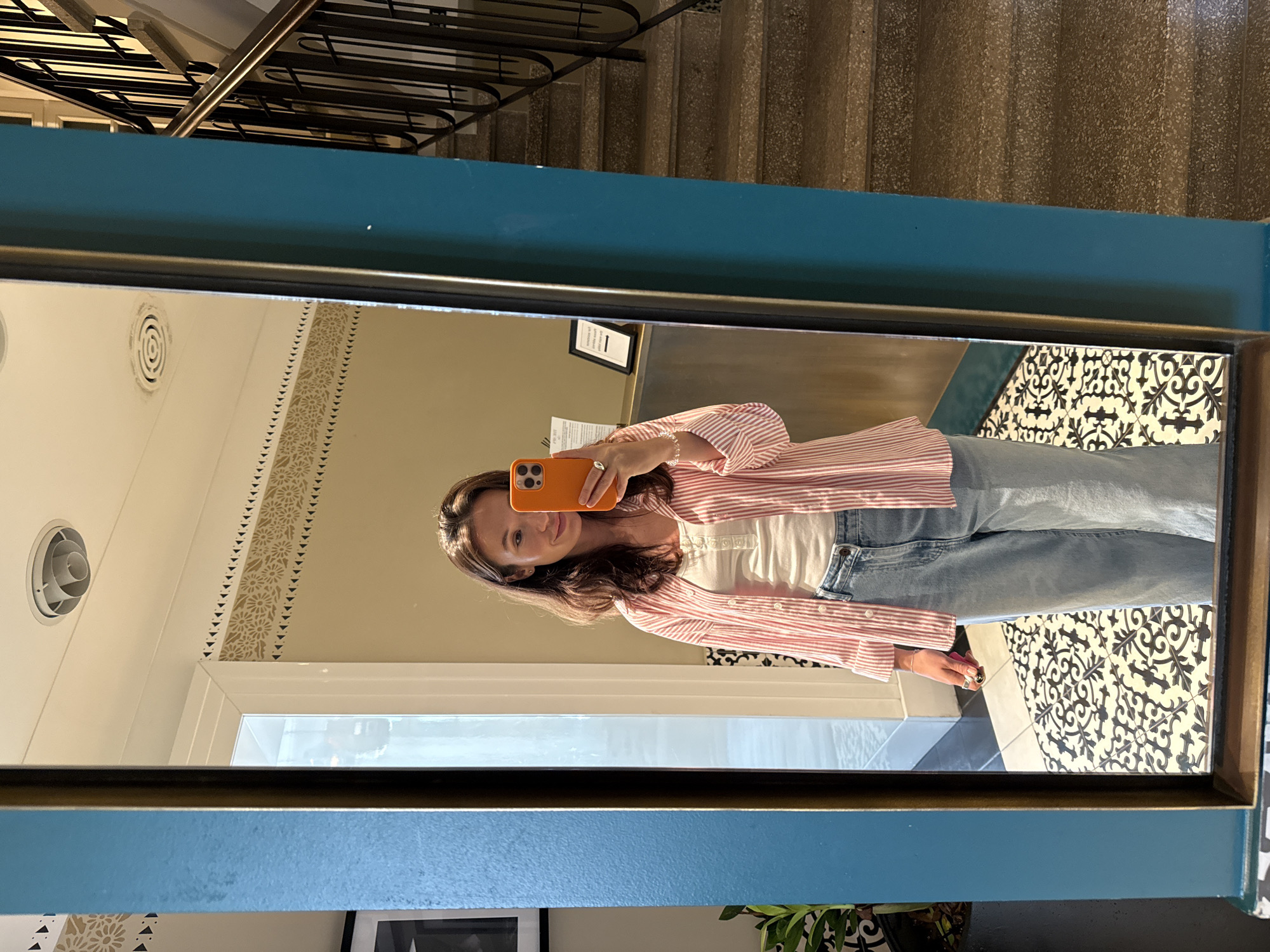 The image is a full-length mirror selfie taken by a young woman indoors. She is standing in front of a mirror with a thick blue frame. She is wearing a white tank top, a pink and white striped button-down shirt, and light-wash wide-leg jeans. She is holding an orange smartphone in her right hand, using it to take the photo. Her left hand is visible at her side, adorned with rings. The background reflected in the mirror shows a tiled floor with a black and white intricate pattern, a counter area, and a staircase with a dark metal railing to the right. A potted plant is visible to the left of the mirror. The woman appears to be in a hotel or a similar establishment. The image does not provide specific visual cues to pinpoint the location within Tel-Aviv, Israel.