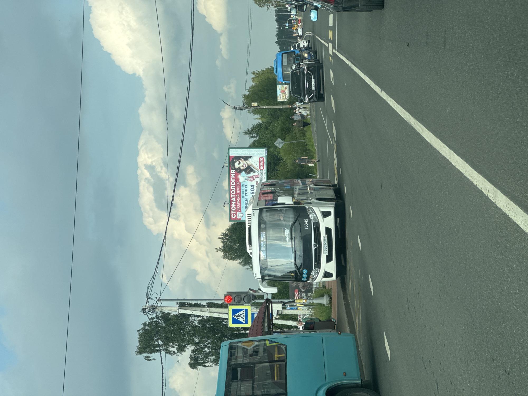 The image depicts a street scene in Novoe Devyatkino, Russia, featuring a pedestrian crossing with a traffic light and a blue bus stopped at a bus stop. A white bus, identified by the number 15342, is also present. Several cars are driving on the road, and a billboard advertising dentistry services is visible in the background. The sky is cloudy, and trees line the side of the road. A few pedestrians are walking along the sidewalk.