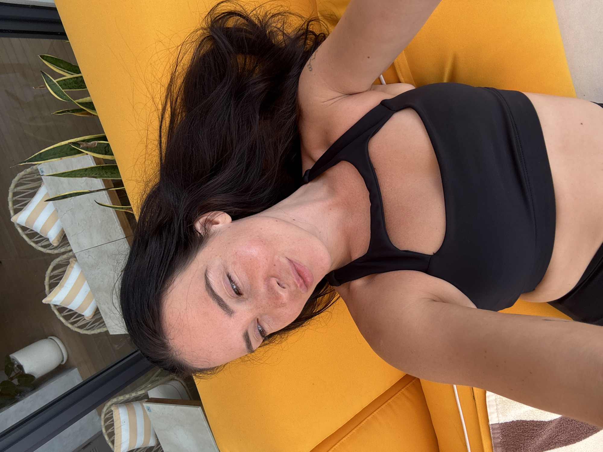 A woman with dark hair and fair skin is lying on a bright orange lounge chair, taking a selfie. She is wearing a black sports bra with a cutout in the front. Her eyes are partially closed, and she has a slight smile on her face. She appears to be outdoors, with a blurry background that suggests a tropical or resort setting with plants and chairs visible through glass. The location is identified as North Kuta, Indonesia.