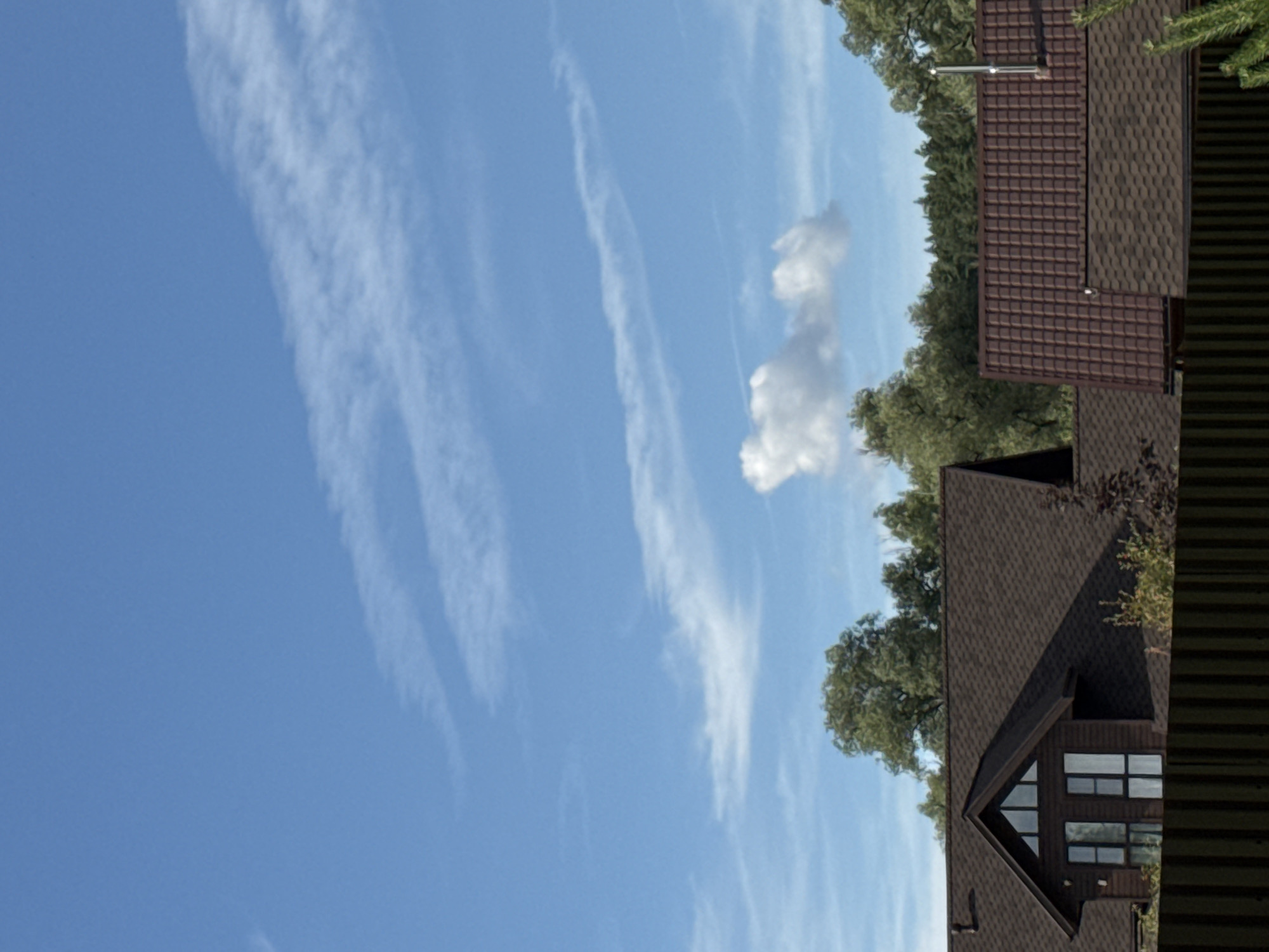 This is a photograph of the sky over a residential area, likely in Serpukhov, Russia. The sky is blue with several wispy white clouds, some appearing as streaks and one larger, puffier cloud. Below the sky are the roofs of several houses, covered in dark brown shingles. Some houses have visible windows, and one has a metal chimney. The tops of green trees are visible behind the houses, suggesting a suburban or rural setting. A dark brown wooden fence runs across the bottom of the image. The scene appears to be taken during daylight hours. There are no people or animals visible in the frame.