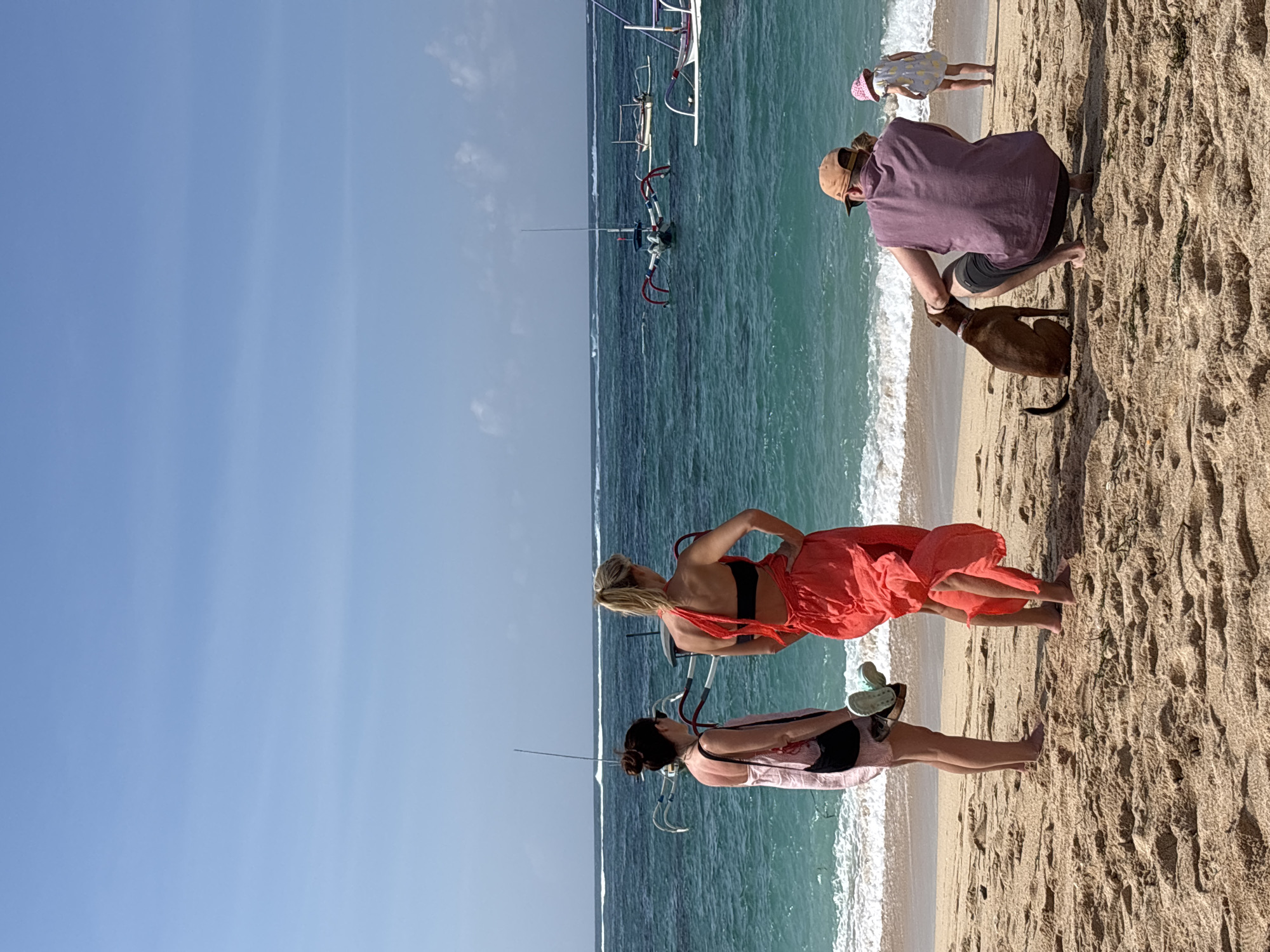 In Kuta Selatan, Indonesia, on a sandy beach under a clear blue sky, two women stand facing the ocean. The woman on the left wears a pink sundress and carries a black bag and light-colored sandals. The woman on the right is dressed in a vibrant red dress and a black bikini top, her hands on her hips. Several traditional outrigger boats are visible on the turquoise water. In the foreground, a man in a purple shirt and a tan hat is crouched on the sand, petting a brown dog. Nearby, a young girl in a yellow dress and pink hat stands facing the water. The beach is composed of light brown sand with some scattered vegetation.