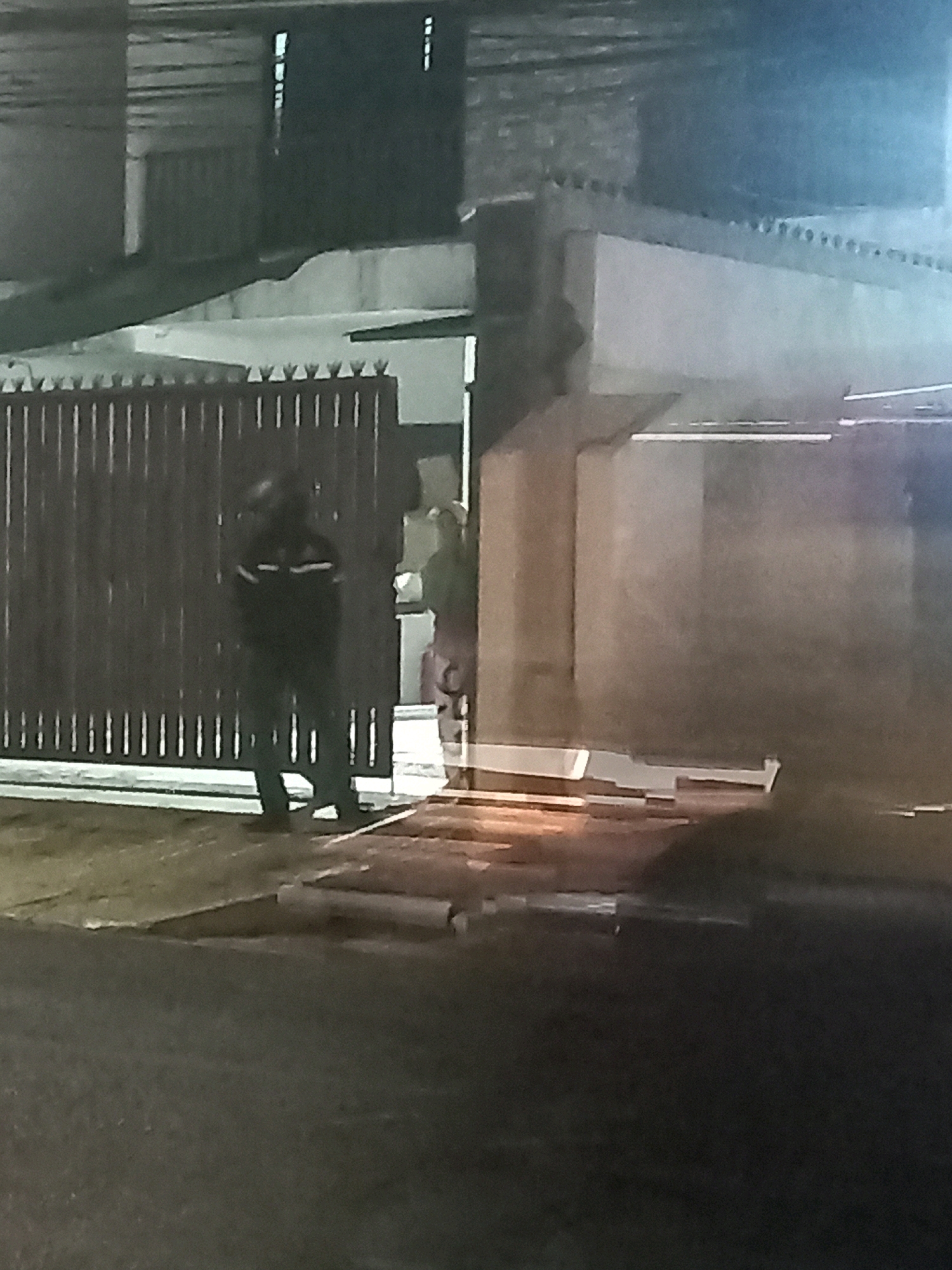 The image, taken at night in South Jakarta, Indonesia, shows two individuals standing in front of a tall, dark metal gate. The gate appears to be a property entrance. The person on the left is standing closer to the gate and is wearing dark clothing with reflective stripes on the chest and arms, suggesting they might be a security guard or a worker on duty. This individual is facing the gate. To the right of this person and slightly further back, another individual is partially visible, also appearing to be standing near the gate. The background shows elements of a building, including walls and a roofline, with some lighting suggesting the area is illuminated. The ground in front of the gate appears to be paved. The overall lighting suggests a low-light condition, typical of nighttime.