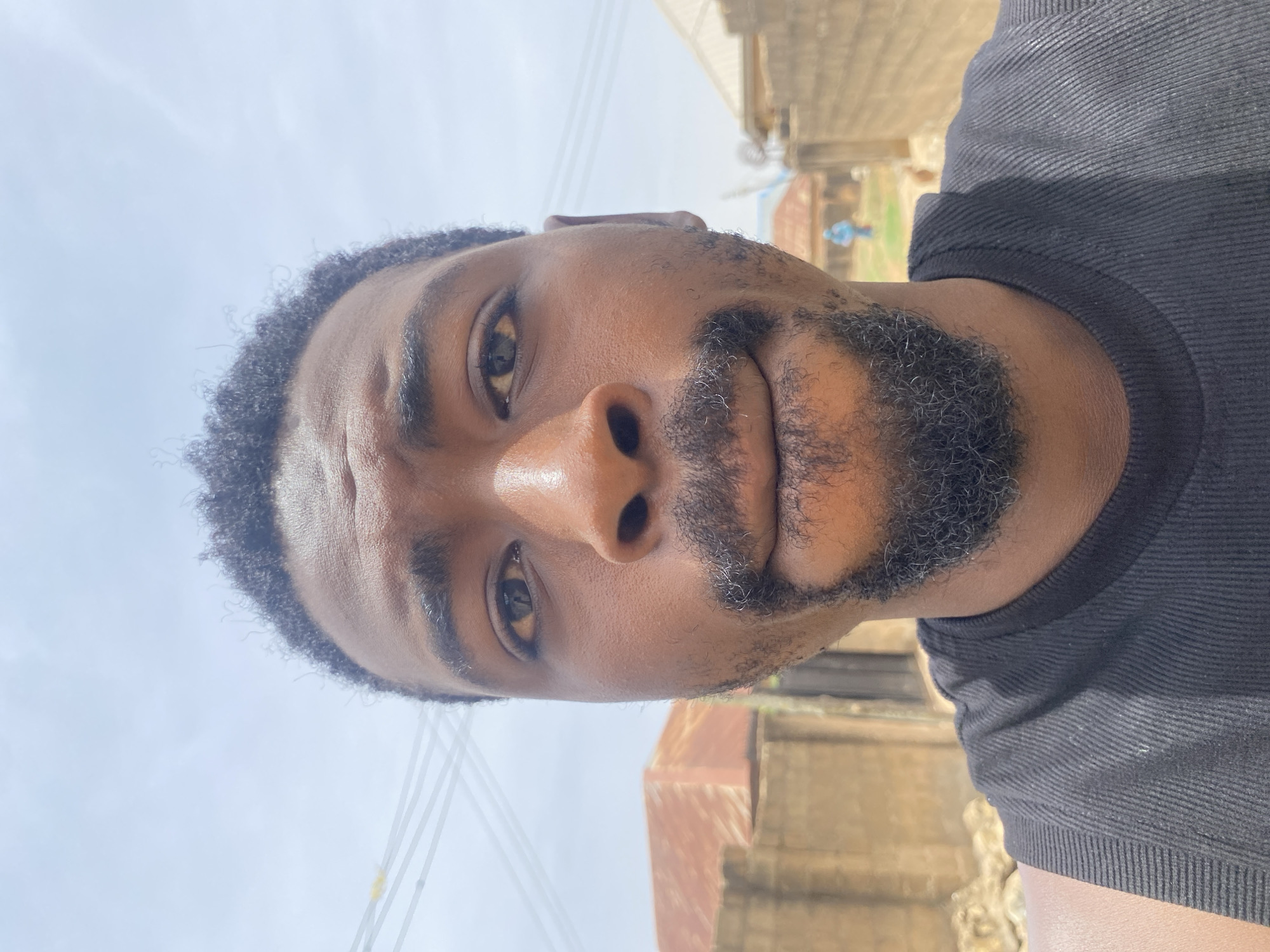 The image is a close-up portrait of a man, initially presented rotated 90 degrees counter-clockwise. When viewed upright, the man is looking directly up towards the camera.
**Subjects:** The primary subject is a man with dark skin, dark, tightly curled hair, and a well-groomed beard and mustache. His dark eyes are open and focused on the camera.
**Objects:** The man is wearing a black, ribbed t-shirt, parts of which are visible on his shoulder and chest.
**Scene/Setting:** The scene is an outdoor setting, characterized by a bright, clear sky that fills much of the background. In the upper right portion of the image (when upright), indistinct architectural structures or buildings with what appear to be simple roofs can be seen. Thin power lines are visible against the sky in the upper left.
**Activity or event taking place:** The image appears to be a selfie or a close-up portrait taken from a low angle, with the subject looking upwards towards the lens.
**Notable details:**
* **Time of day:** The bright, light blue sky indicates clear daytime conditions, likely midday or early afternoon.
* **Weather:** The weather appears clear and sunny, with no clouds visible.
* **Location cues:** The simple background structures and visible power lines are consistent with an outdoor setting in a developing area, aligning with the provided location context of Barakin Akawo, Nigeria.
* **Emotions:** The man's expression is neutral and composed, with a direct gaze.
* **Visible text:** There is no visible text in the image.