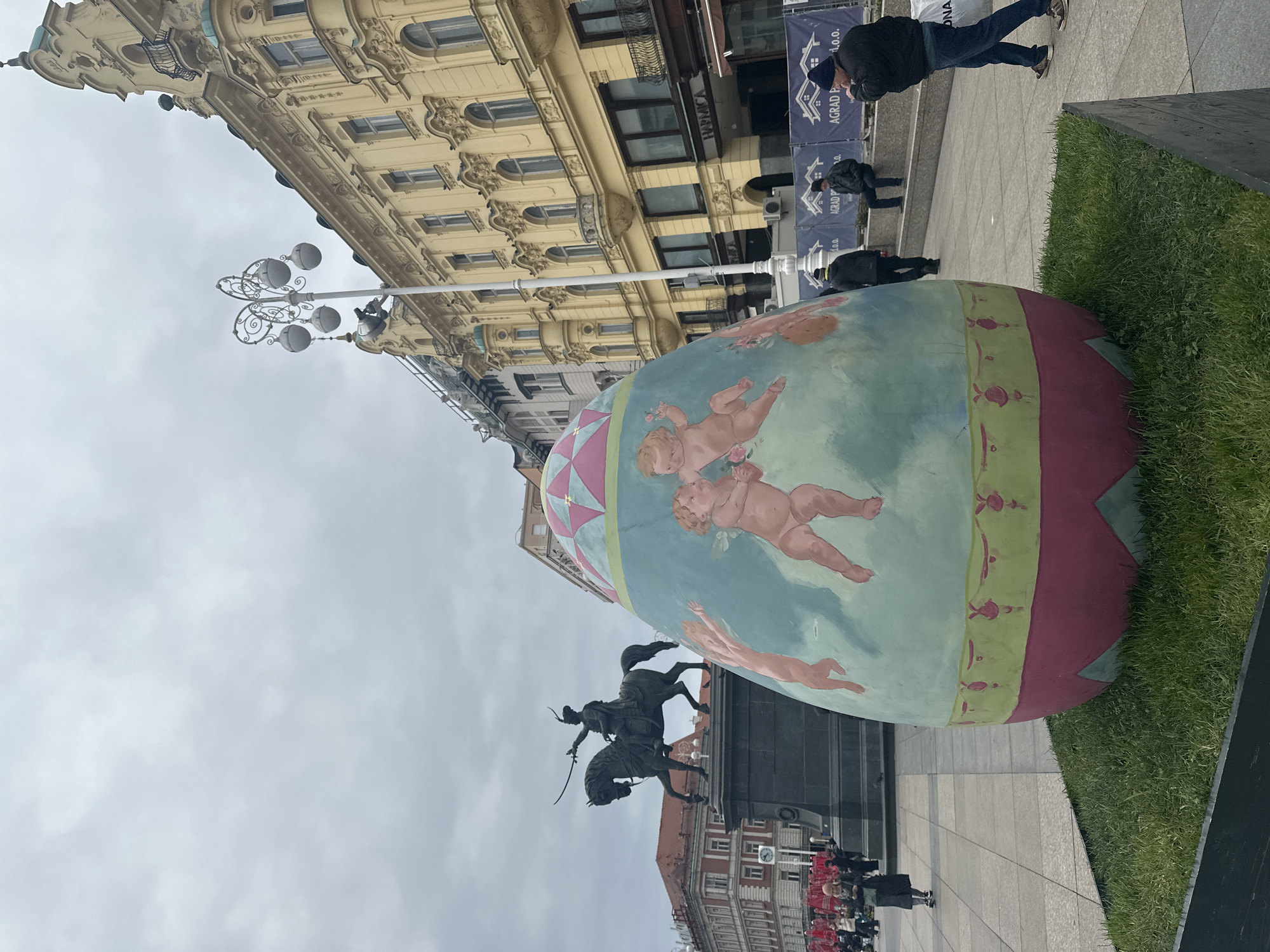 The image shows a street scene in Zagreb, Croatia, featuring a large, decorated Easter egg prominently displayed near a statue. The egg is painted with images of cherubs and geometric patterns, standing on the cobblestone sidewalk. Behind the egg stands the equestrian statue of Josip Jelačić on Trg bana Jelačića, the city’s central square. Several people are visible in the background, some walking. A light post is also visible. The buildings surrounding the square are in a classic European architectural style. The sky is overcast, indicating diffused daylight.