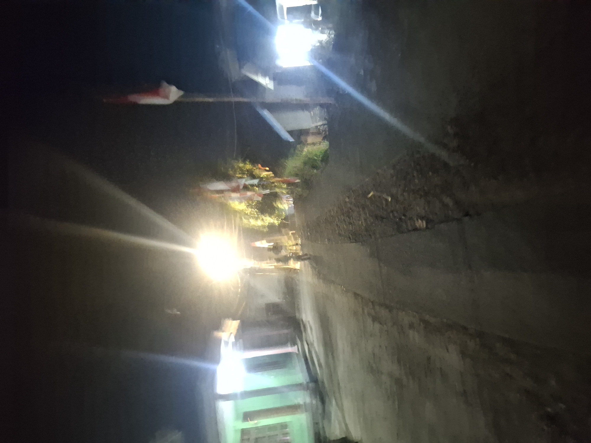 The image depicts a dimly lit street scene at night in Muncar, Indonesia. A bright street light illuminates the asphalt road, casting long shadows and creating lens flares. On the left side of the road, a building with green walls and white columns is visible. To the right of the road, several small buildings with peaked roofs are present, one of which is brightly lit from within. Along the right side of the road, a row of Indonesian flags, displaying red and white colors, are planted on poles, suggesting a national celebration or event. In the middle of the road, a person on a bicycle is riding away from the camera. The overall atmosphere is somber due to the low light conditions, but the flags and the illuminated buildings suggest a community gathering or a festive occasion. The ground appears to be a mixture of asphalt and dirt.