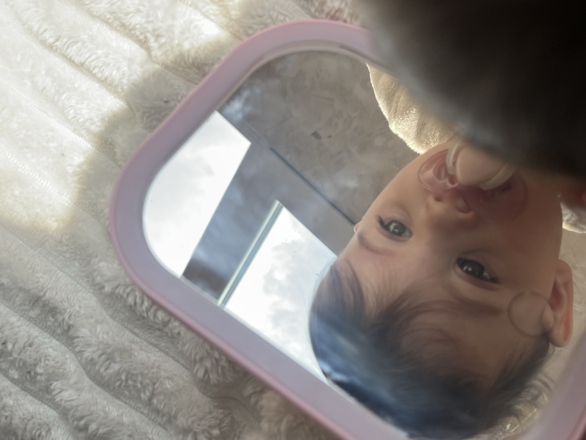A light-skinned infant with dark hair and eyes is depicted in a reflection within a pink-framed mirror. The baby has a clear or light pink pacifier in its mouth and is looking forward. The mirror rests on a textured, white, fuzzy surface with visible quilting stitches. The reflection within the mirror shows a bright window with sky and clouds, along with a darker interior surface. A dark, indistinct object partially overlays the lower right portion of the mirror and the infant's face. The scene is illuminated by natural light, occurring indoors in Elena, Bulgaria.