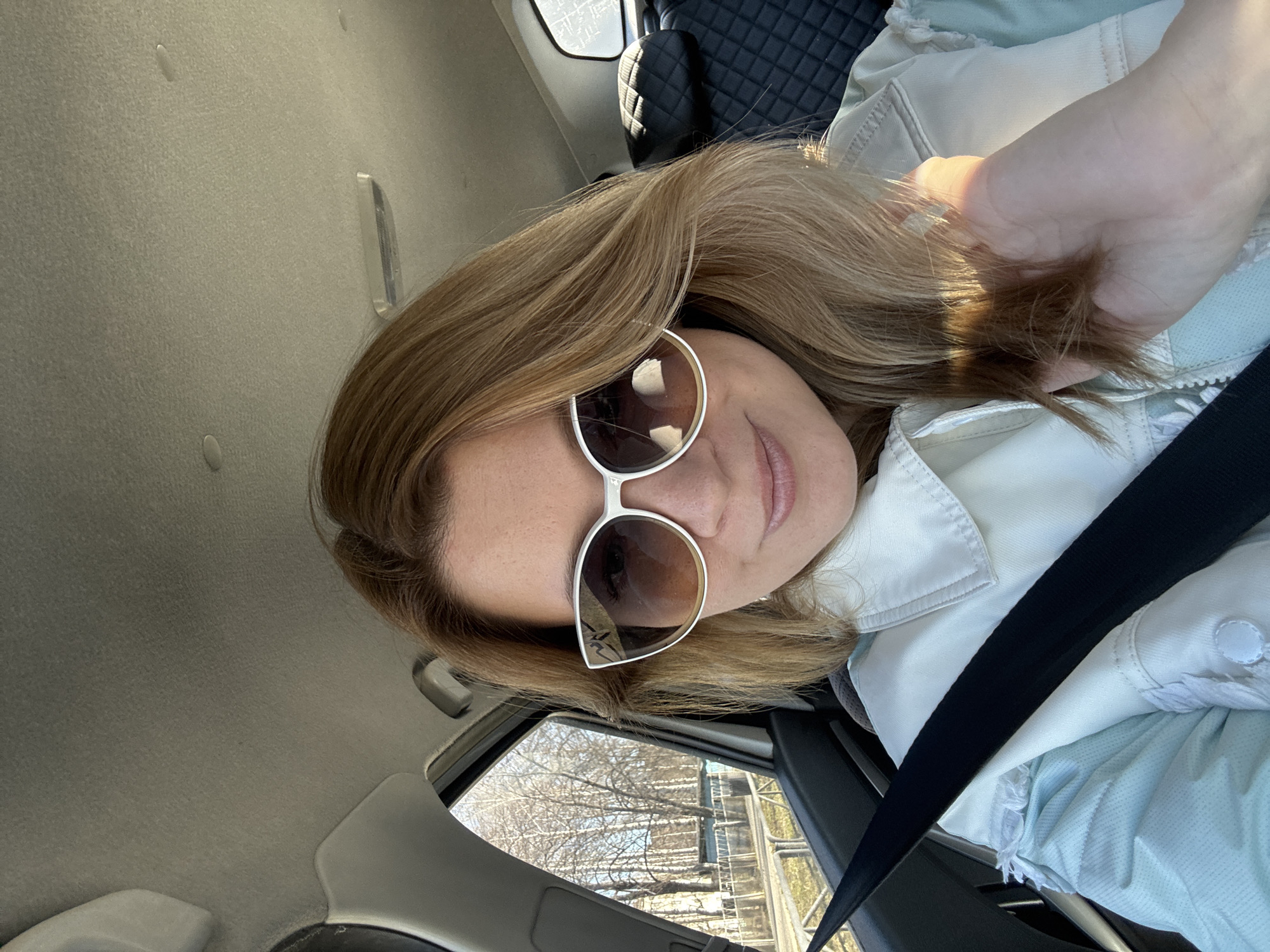 A woman is sitting in the driver's seat of a car, wearing sunglasses and a white jacket. She is wearing a seatbelt. Her left hand is holding a clump of her own hair. Through the car window, bare trees and a metal railing or bridge structure are visible. The location appears to be outdoors, potentially on a road or bridge in Chaikovsky, Russia based on the presence of trees, a structure resembling a guardrail or bridge, and the urban surroundings suggested by the car's interior.