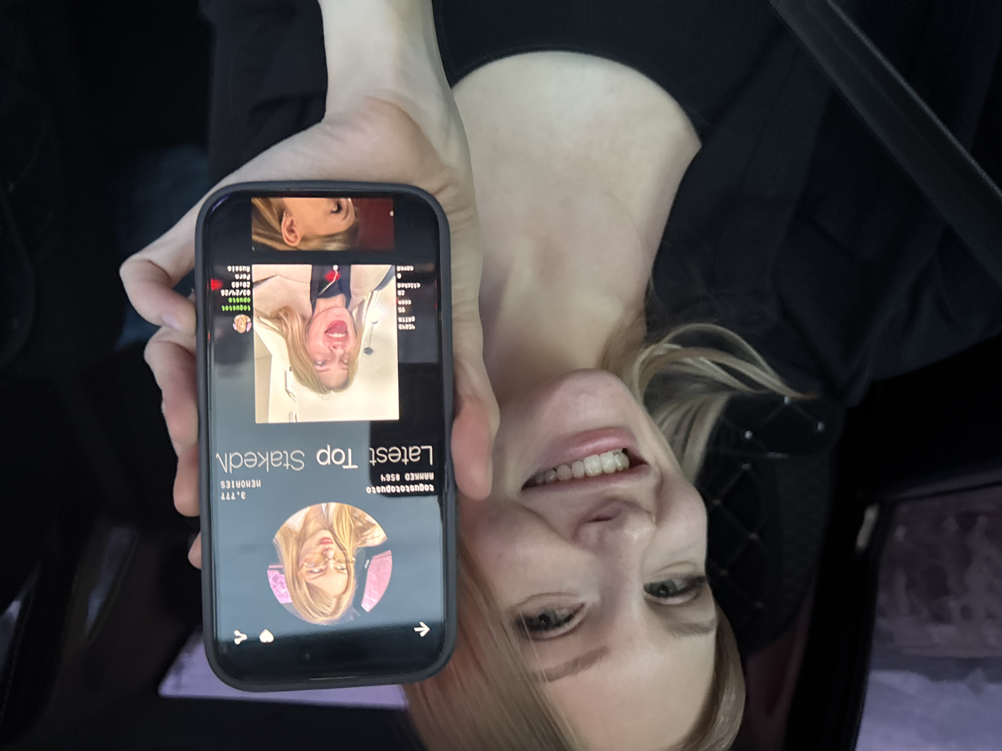 A woman in what appears to be a car holds up her smartphone displaying a social media profile. The displayed profile on the phone features another woman with blonde hair. Text on the profile includes "Latest Top Staked" and mentions "togustotopusto", "RANKED #564", "3.777 MEMORIES", and information such as "clicked 20", "03/24/20", "20:03", and "Russia". It also includes "Perm".