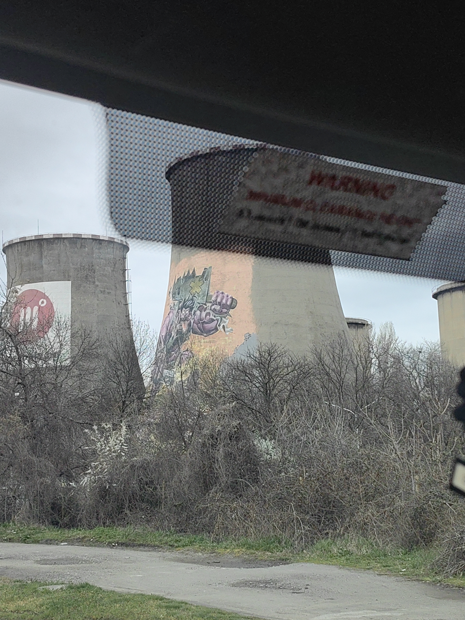 A photograph shows cooling towers, possibly part of a power plant, located in Sofia, Bulgaria. The towers are painted with graffiti art. One tower has a circular red design on a white background. The other has a purple, green, and pink monster-like figure. Bare trees and bushes surround the base of the towers. A road and grassy area are in the foreground. The photograph is taken from inside a vehicle; a section of the visor with a "WARNING" label is visible at the top of the image. The sky is overcast.