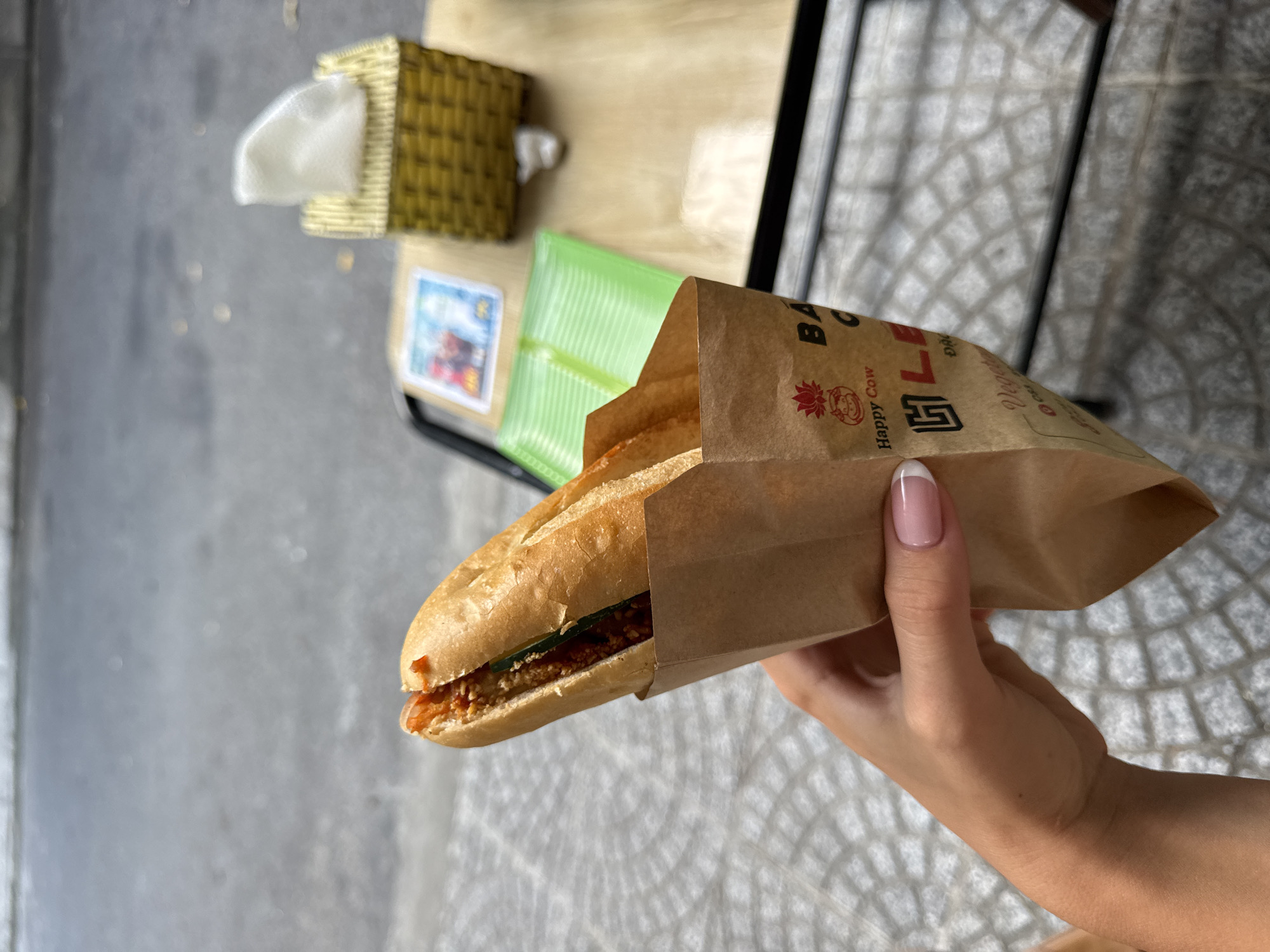 An eye-level, medium shot shows a baguette-style sandwich in a brown paper bag held by a fair-skinned human hand with pink and white manicured nails. The sandwich appears to be stuffed with meat and lettuce. The brown paper bag is printed with the image of a cartoon cow. The bag is printed with "Bà LE," a restaurant located in the city of Đà Nẵng, Vietnam.
In the background, out of focus, is a sidewalk and a small table with a woven tissue box. The sidewalk is patterned with a light grey lattice design.
