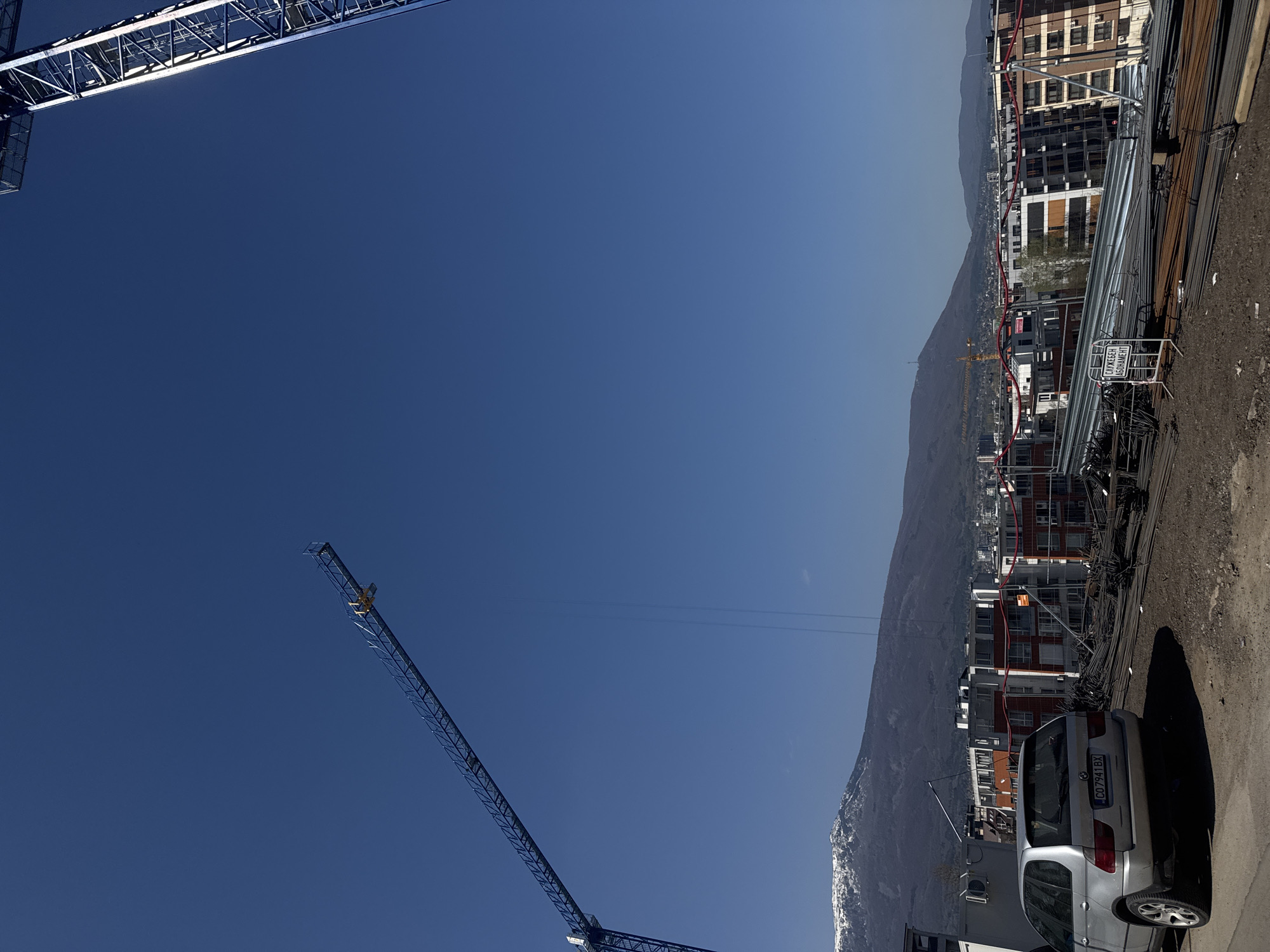 This image captures a vibrant construction scene in Sofia, Bulgaria, under a clear, bright blue sky. Two large tower cranes are prominent, with one partially visible in the upper left corner and another spanning diagonally from the lower left towards the middle right, indicating active development.
On the right side of the frame, several multi-story buildings, some appearing new or still under construction, line what seems to be a road or access area. Piles of metal rebar and other building materials are stacked along the ground next to these structures. Amidst these materials, a white sign with black Cyrillic text is visible, reading "СЛУЖЕБЕН АБОНАМЕНТ" (SLUZHEBEN ABONAMENT), which translates to "SERVICE SUBSCRIPTION" or "OFFICIAL SUBSCRIPTION."
In the foreground on the right, a silver SUV, identified as a BMW X5, is parked. Its Bulgarian license plate, "CO 7941 BX," explicitly confirms its Sofia registration ("CO").
The background is dominated by a majestic, expansive mountain range, partially snow-capped, strongly suggesting the iconic Vitosha Mountain that borders the city of Sofia. A smaller yellow construction crane can be faintly seen in the distance near the mountain's base.
The time of day appears to be mid-morning or early afternoon, given the bright, sunny weather and the absence of shadows that would indicate early morning or late afternoon. The overall atmosphere conveys ongoing urban expansion and infrastructure development within a scenic natural setting. No individuals are visible in the photograph.