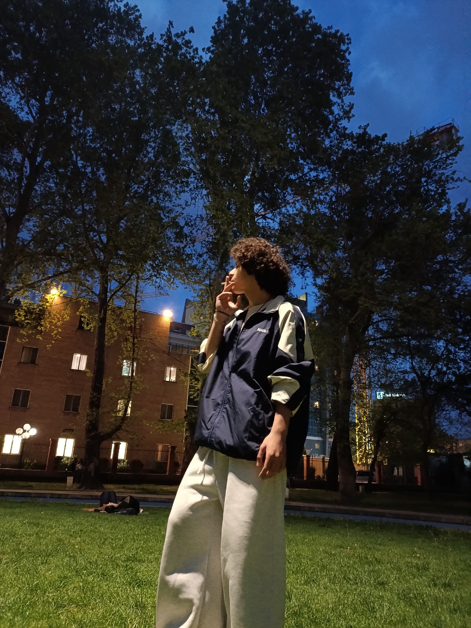 A person with curly hair wearing a navy blue and white jacket and light gray sweatpants stands on a grassy lawn. The person holds something to their mouth, possibly smoking. In the background are trees, a brick building with lit windows, and a building with a construction crane behind it. In the foreground, on the grass, someone lies prone. The scene appears to be lit by streetlights and takes place at night. It is likely in Yerevan, Armenia.