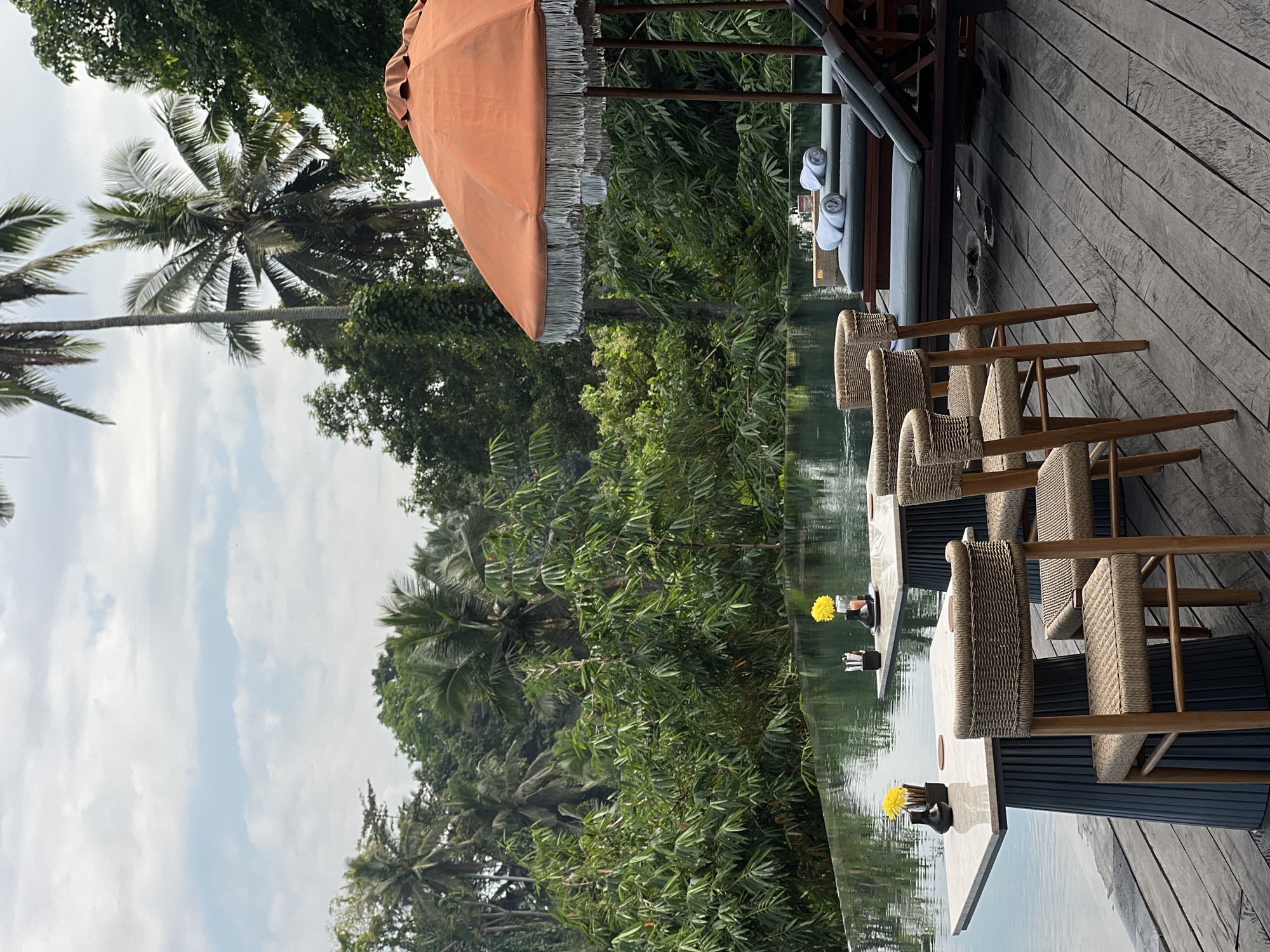 The image depicts an outdoor seating area at a resort, likely overlooking a pool and lush tropical foliage in the Ubud District of Indonesia. The foreground features a wooden deck with several wicker chairs and marble-topped tables set for dining. Two tables are visible, each adorned with a single yellow flower in a small black vase and cutlery holders. Beyond the tables, an infinity pool reflects the overcast sky and the dense greenery of palm trees and other tropical plants.
To the right of the pool, a set of lounge chairs are arranged under a large, orange fringed umbrella. These chairs are covered with dark cushions and topped with neatly rolled white towels. The surrounding environment is characterized by vibrant green vegetation, including tall palm trees characteristic of the region. The atmosphere suggests a serene and luxurious setting, typical of high-end accommodations in Ubud, known for its natural beauty and tranquil ambiance. There are no people visible in the image, implying the scene is either empty or captured during a quiet moment.