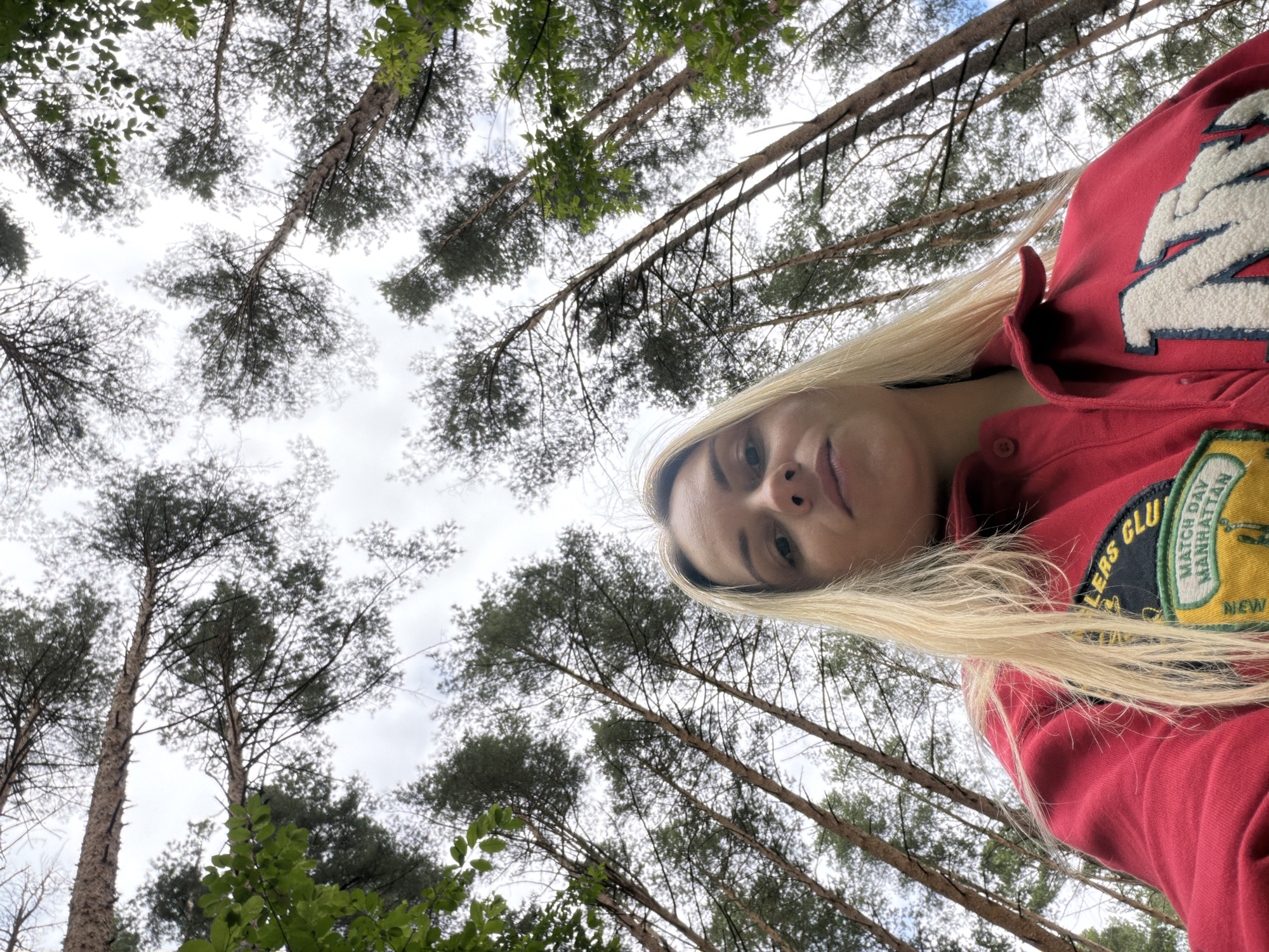 An extreme low-angle, medium shot captures a young Caucasian woman with blonde hair, looking up at the camera. She is wearing a red hooded jacket with patches on the chest. The background consists of a canopy of tall trees, their trunks rising towards a cloudy sky. The image appears to be taken outdoors in a forest setting. There is no information available regarding the location in Votkinsk, Russia.