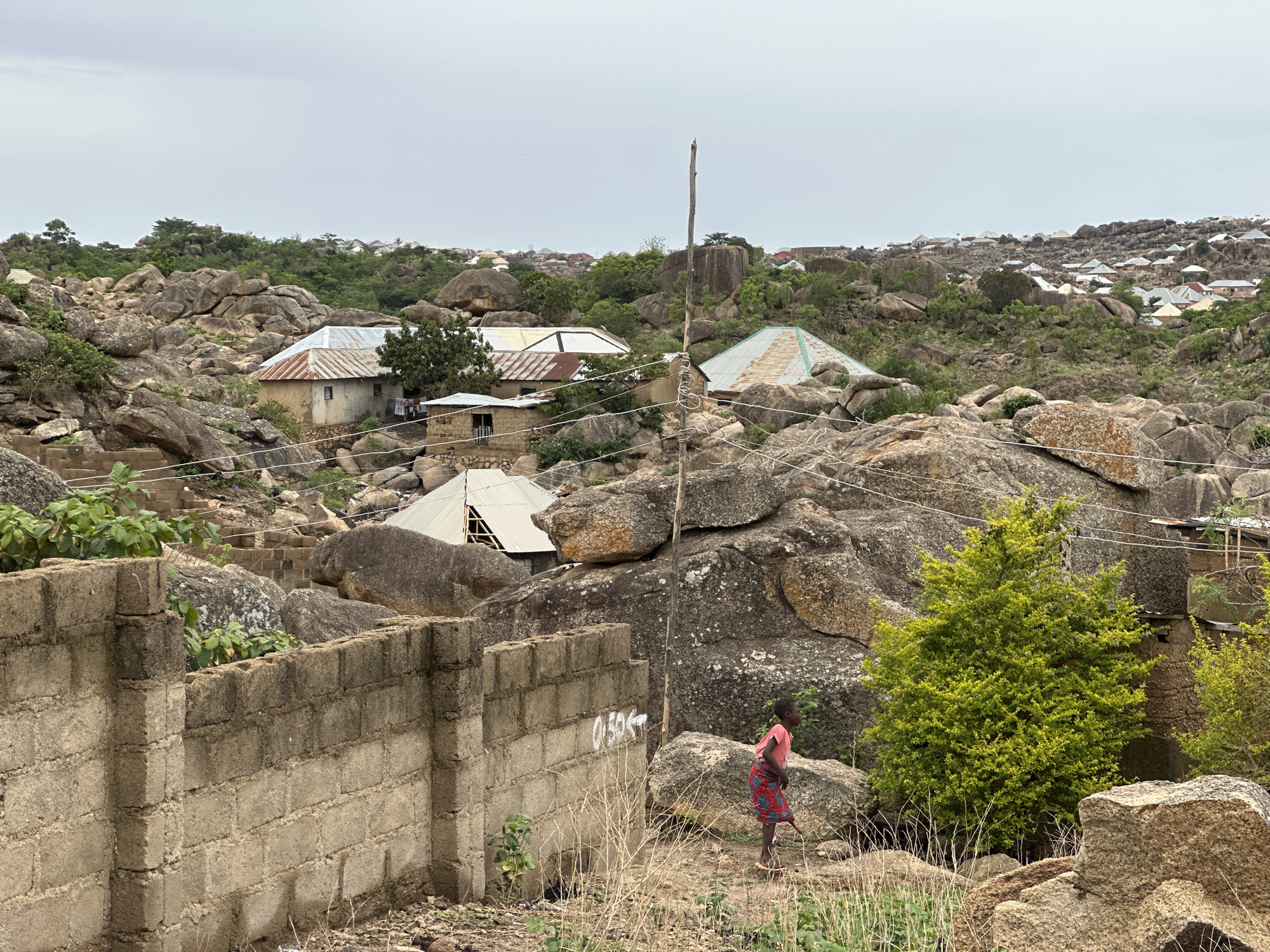 The scene depicts a landscape in Jos, Nigeria, characterized by a mix of natural and human-made elements. Prominent features include large, weathered granite boulders scattered throughout the area, interspersed with low-lying vegetation and small trees. Several single-story buildings with corrugated metal roofs are nestled among the rocks, suggesting a settlement integrated into the natural terrain. In the foreground, a roughly constructed concrete block wall stands, partially obscuring the view. A young person wearing a pink shirt and patterned skirt walks along a path near the wall and rocks. Electrical wires are strung across the landscape. The sky is overcast and light gray.