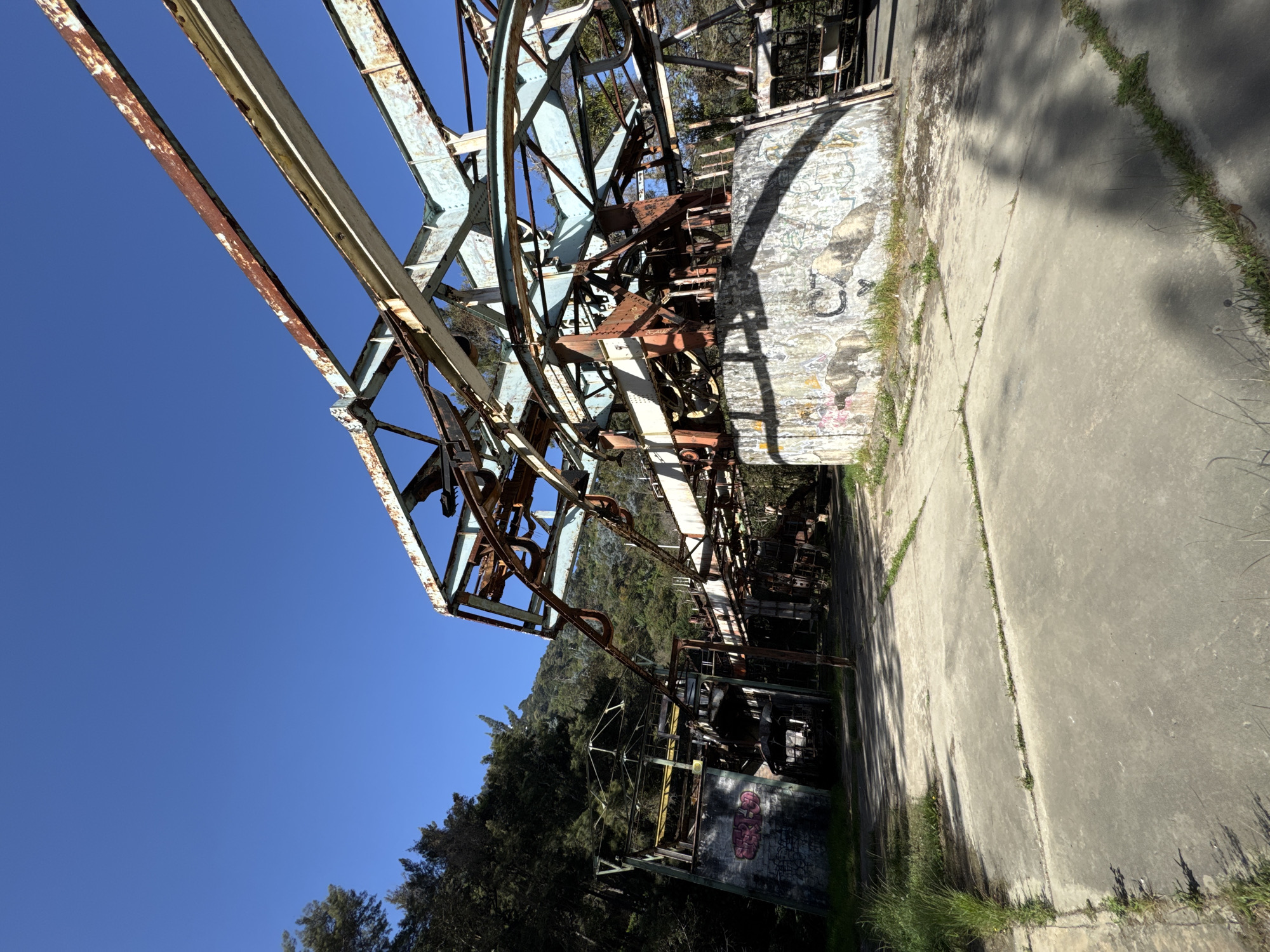 The image shows a dilapidated, rusty metal structure, possibly part of an abandoned cable car system, set against a clear blue sky and a backdrop of green trees. The structure's steel frame is heavily corroded with significant rust, suggesting long-term disuse and exposure to the elements. The structure is located in San Antonio de Galipán, Venezuela. It is situated next to a concrete pathway or platform. The image is rotated counter-clockwise. A concrete wall adorned with graffiti is visible.