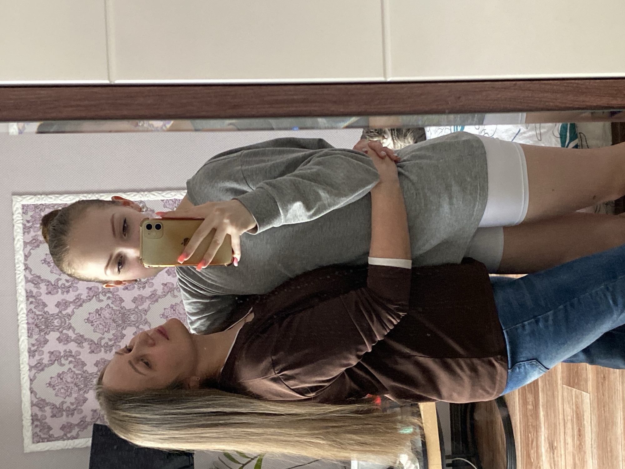 An indoor mirror selfie captures two females. The image is oriented vertically with the subjects appearing as reflections. In the foreground reflection, an adult woman with long, straight blonde hair, wearing a dark brown long-sleeved top and blue jeans, looks upwards and slightly to the right. Behind her, a younger female, possibly a teenager, with light brown hair pulled into a bun, is taking the photo. She is wearing a grey long-sleeved top and white shorts or underwear. She holds a light brown/gold smartphone with a multi-lens camera, her pink-painted fingernails visible. Her gaze is directed towards the phone's screen.
The setting appears to be a bedroom or living space. Visible in the real-world background (as seen through the mirror) is a wall covered in light purple wallpaper with a white floral pattern, framed in white. A wooden floor is visible towards the bottom right. The mirror itself has a dark brown frame at the top, above which is a plain light-colored ceiling or wall. Reflected in the mirror behind the younger girl's legs, parts of a bed with a white sheet and a patterned comforter are visible. The overall mood is casual. No specific text is visible in the image.