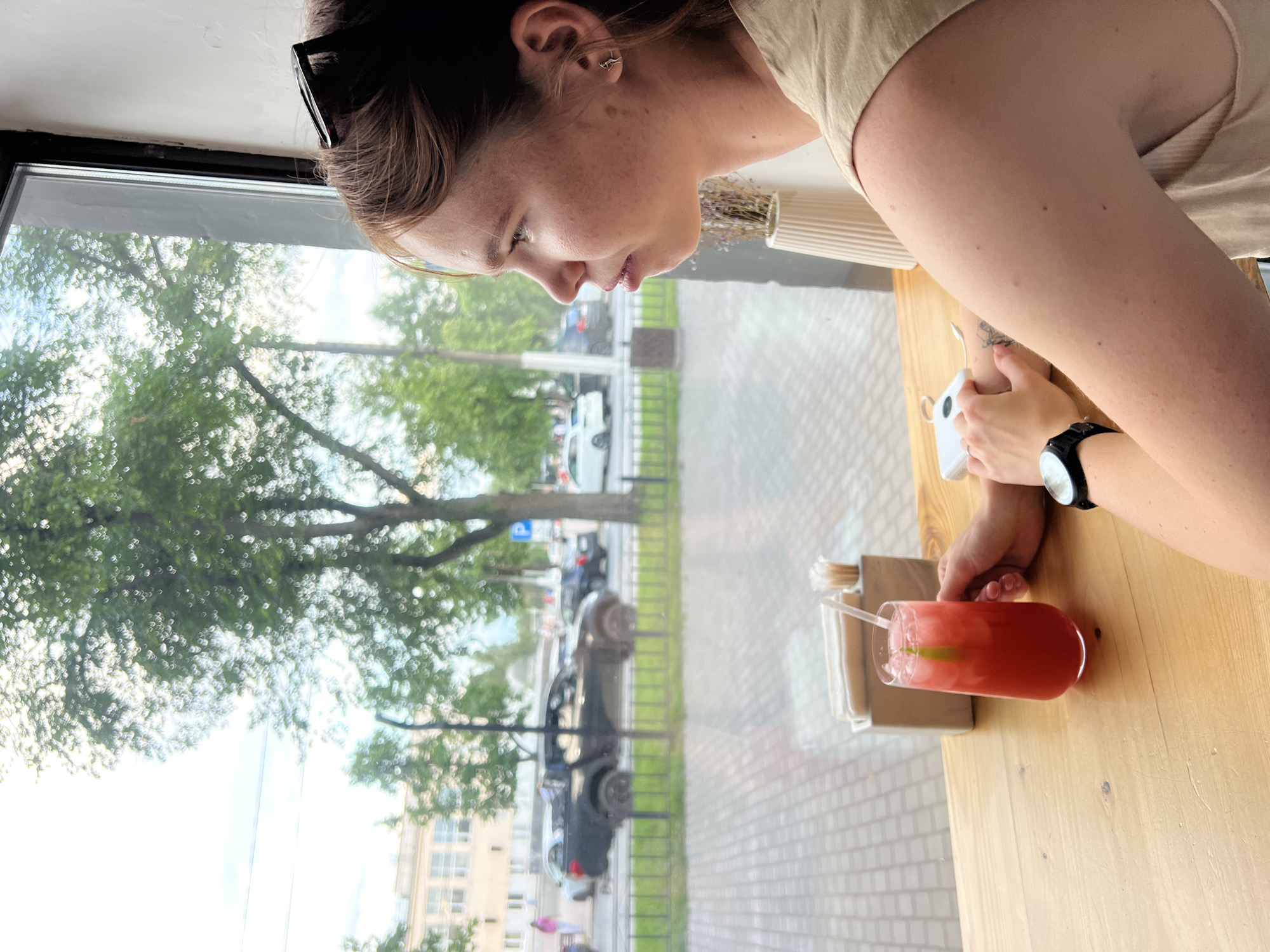 A young woman with freckles and light brown hair is sitting at a wooden table by a window, looking down at a pink drink with ice and a lime slice in a clear glass. She is wearing a light beige t-shirt and a black watch on her left wrist. Sunglasses are perched on top of her head. Her right hand is resting on the table near the drink. Through the window, trees with green leaves are visible, along with a street with parked cars and a sidewalk. The time of day appears to be daytime, with bright natural light. The scene suggests a casual setting, possibly a cafe or restaurant, in Chaikovsky, Russia. The woman's expression is neutral, and there is no visible text in the image.