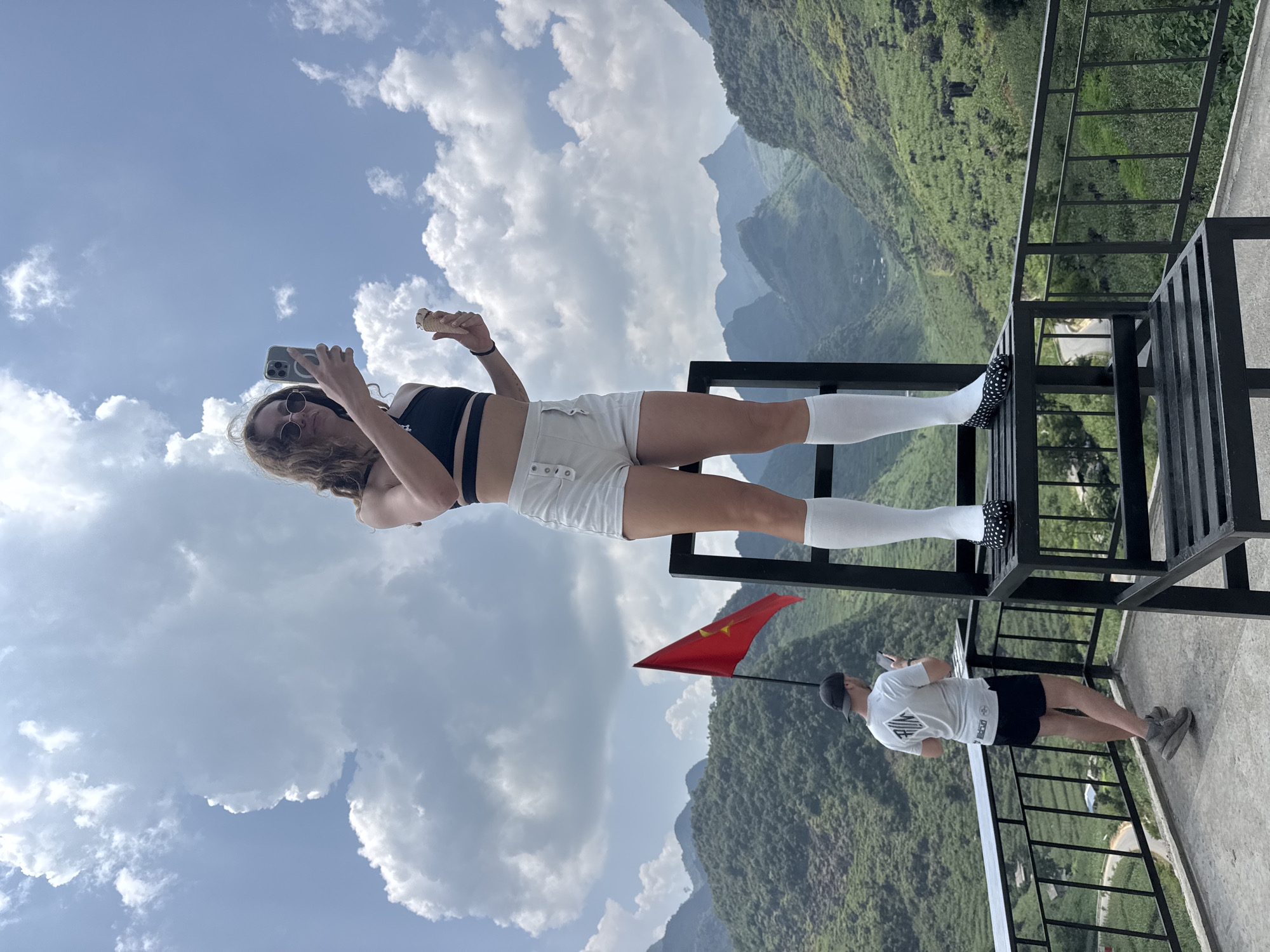 A woman, with her center of gravity supported on a metal structure that overlooks mountainous terrain, holds a smartphone in one hand and an ice cream cone in the other. A Vietnamese flag stands adjacent to a man who is holding a smartphone, also adjacent to the mountainous terrain. The city is Lũng Phìn Commune, Vietnam. The sky is partly cloudy. The woman is wearing white shorts, white knee-high socks, and a black top. The man is wearing a white t-shirt, black shorts, and a black baseball cap.