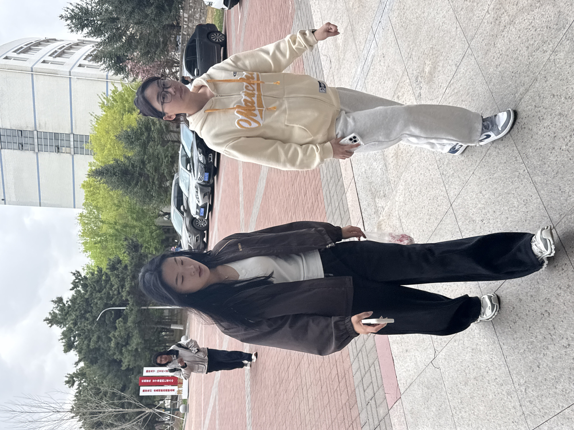 Here is a description of the image:
Two young individuals are captured standing outside in what appears to be a university or college campus in Zhongtun, China. The first individual, on the upper side of the frame, is wearing glasses, a beige zippered hoodie with the word "March" written on it, and grey sweatpants, and sneakers. The second individual, on the lower part of the frame, has long dark hair, a brown bomber jacket over a white top, black pants, and sneakers. Both appear to be holding phones.
The setting shows elements of an academic environment with a building in the background, along with trees and cars parked along the side. A sign displaying Chinese characters suggests local regulations or campus guidelines. The brick-paved ground indicates a pedestrian area.