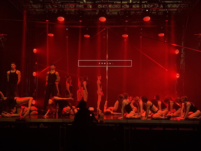 The image captures a stage performance illuminated by intense red lighting within an event venue in Moscow, Russia. The stage features a prominent, tall chrome pole positioned centrally. On the left side of the stage, two male performers are visible; both wear dark tank tops and dark pants. The male performer further right has visible tattoos on his arms. To the right of the pole, a group of approximately 10-15 female performers in dark, form-fitting athletic wear are positioned on a reflective stage surface, primarily in kneeling or crouching poses. The background consists of a dark environment with visible stage trussing and multiple red LED spotlights casting beams through what appears to be stage fog or haze. A large screen or projection in the background displays the white Russian word "ТРИБУНА" (Tribuna) within a white rectangular border. In the immediate foreground, a silhouetted figure is partially visible, facing the stage.