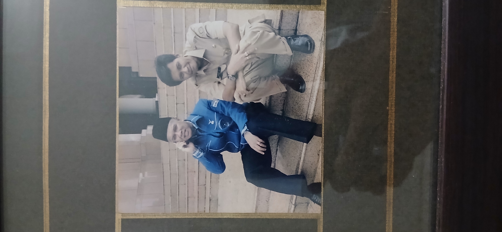 Two Indonesian men are seated on concrete steps outdoors. The man on the left wears a black peci hat, a blue jacket with white trim and logos, and dark pants. He is holding a mobile phone to his right ear. The man on the right wears a light brown uniform with epaulets and dark pants. He is leaning forward, resting his forearms on his knees, and smiling. The background consists of more concrete steps and a building facade. The image appears to be a framed photograph, with a dark border and gold accents. Based on the attire of the man in uniform and common cultural cues, this photo likely depicts individuals associated with Indonesian government or civil service. Without further context, pinpointing the exact location within Samarinda, Indonesia, is not possible.
