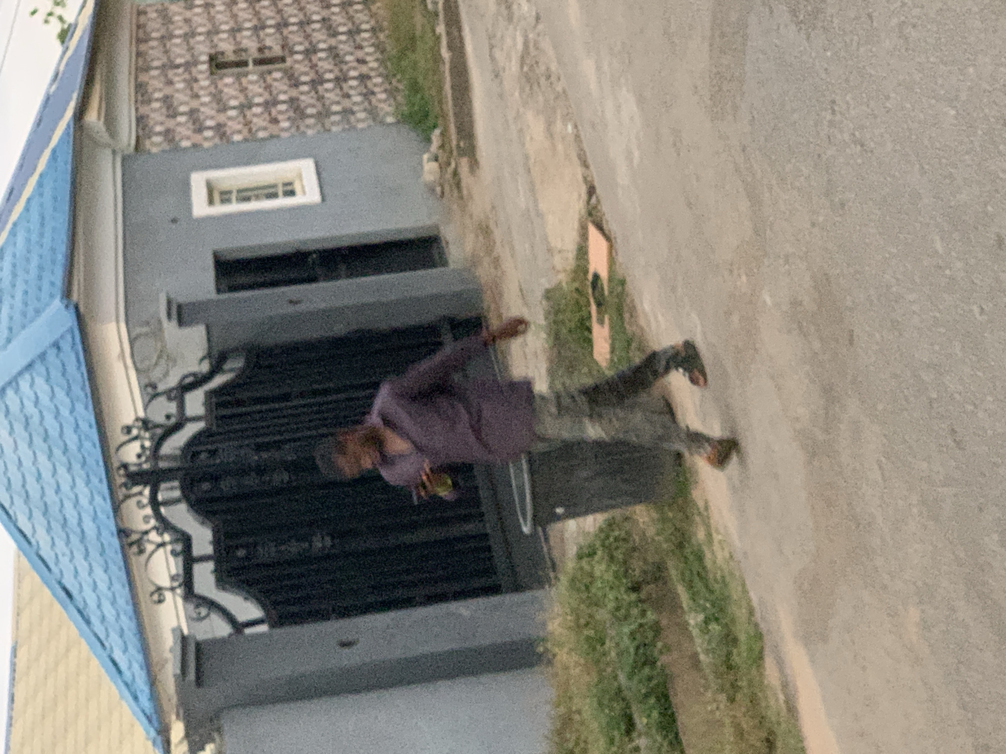 A man wearing a purple long-sleeve shirt and dark pants is walking on a sidewalk in front of a building. The building has a dark iron gate, a gray exterior, and a blue-tiled roof. A window is visible on the upper floor. The location is the city of Abuja, Nigeria.