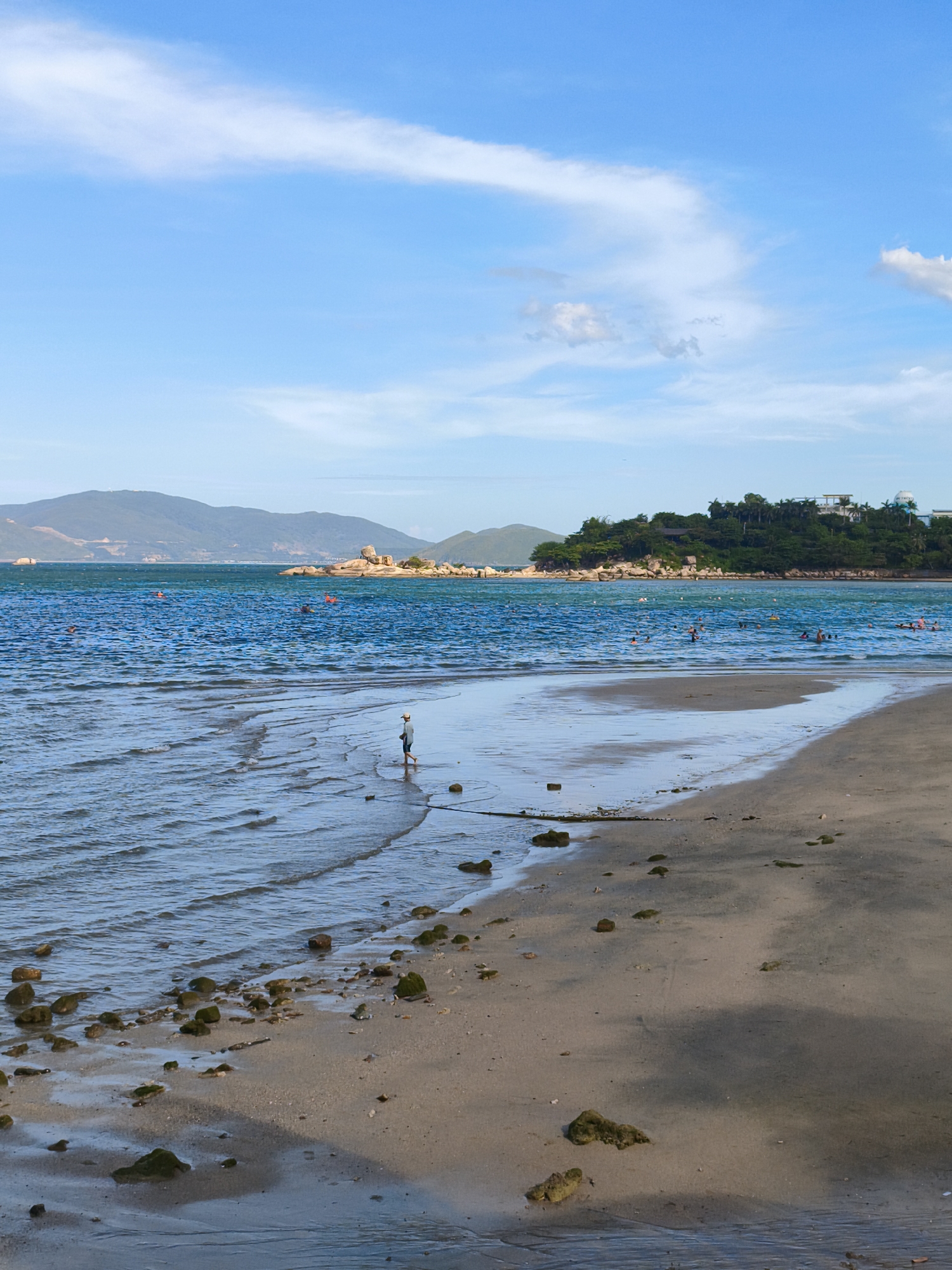 The image depicts a serene coastal scene in Nha Trang, Vietnam, likely on one of its popular beaches. The foreground is dominated by a sandy beach with scattered rocks and pebbles. The tide is out, revealing ripples and shallow water extending towards the sea. In the middle ground, a lone individual, appearing to be a young person wearing a hat and dark clothing, walks near the water's edge. The sea itself is a vibrant blue, with small waves gently lapping the shore. Further out, several people are visible swimming and enjoying the water, marked by orange buoys that likely denote a swimming area or safety zone. In the background, a lush green island covered in trees and buildings rises from the sea, with a mountainous landscape stretching beyond it under a bright blue sky dotted with wispy white clouds. The overall impression is of a tranquil day at the beach, with a mix of natural beauty and human activity.