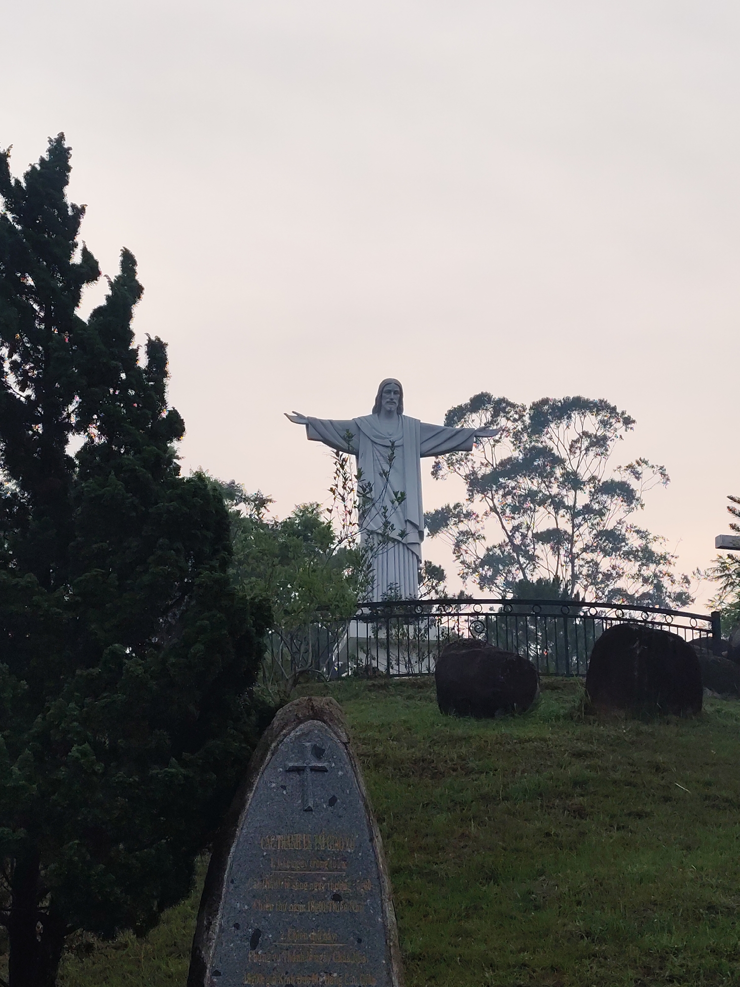 The image presents a tranquil outdoor scene featuring a large statue of Jesus Christ with outstretched arms, standing prominently on a grassy hill. The statue, rendered in a light grey or white material, depicts Jesus in a flowing robe, reminiscent of the Christ the Redeemer style, and is positioned behind a decorative black metal fence.
In the immediate foreground on the left, a tall, dark green coniferous tree dominates the lower-left section of the frame, its dense foliage providing a stark contrast to the brighter background. On the right side of the foreground, two large, dark, rounded boulders are settled on the green, manicured grass. In the bottom-left corner, a dark, triangular stone monument or plaque is visible. It features a carved cross at its top and contains faint, inscribed Vietnamese text, which includes words like "VIỆT NAM" and numbers such as "1800", suggesting it may be a historical marker or dedication.
The background consists of various trees with lighter, more scattered foliage, set against a uniformly light and muted sky. The soft, diffused light indicates either an overcast day, early morning, or late afternoon, contributing to a serene and reflective atmosphere. The scene captures a moment of stillness, emphasizing the monument itself and its natural surroundings. No specific activity or event is taking place beyond the static presence of the religious monument. This setting is located in Lang Biang, Da Lat, Vietnam.