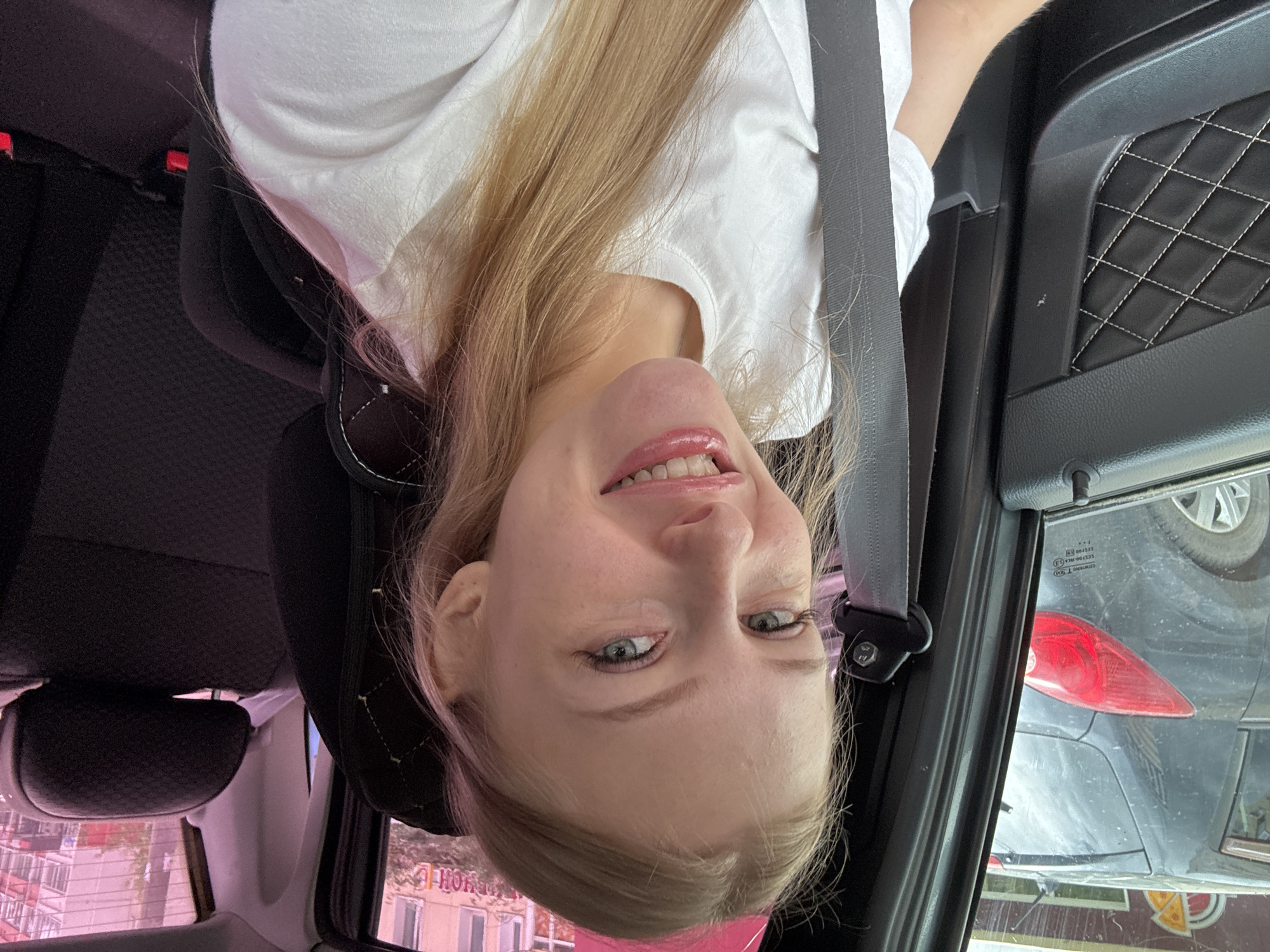 A young Caucasian woman with blonde hair and blue eyes is inside a car, looking directly at the camera and smiling. She is wearing a white t-shirt. A gray seatbelt is visible across her chest. The interior of the car shows a black quilted door panel on the left and black car seats in the background. Through the car window on the left, another car is visible, and beyond that, a building with a sign that appears to say "Pizza" in a circular logo. Through the window on the right, a building with multiple floors and windows is visible, along with trees. The overall lighting suggests it is daytime. The location is identified as Perm, Russia, based on the visual cues, though this cannot be definitively confirmed by the image content alone.