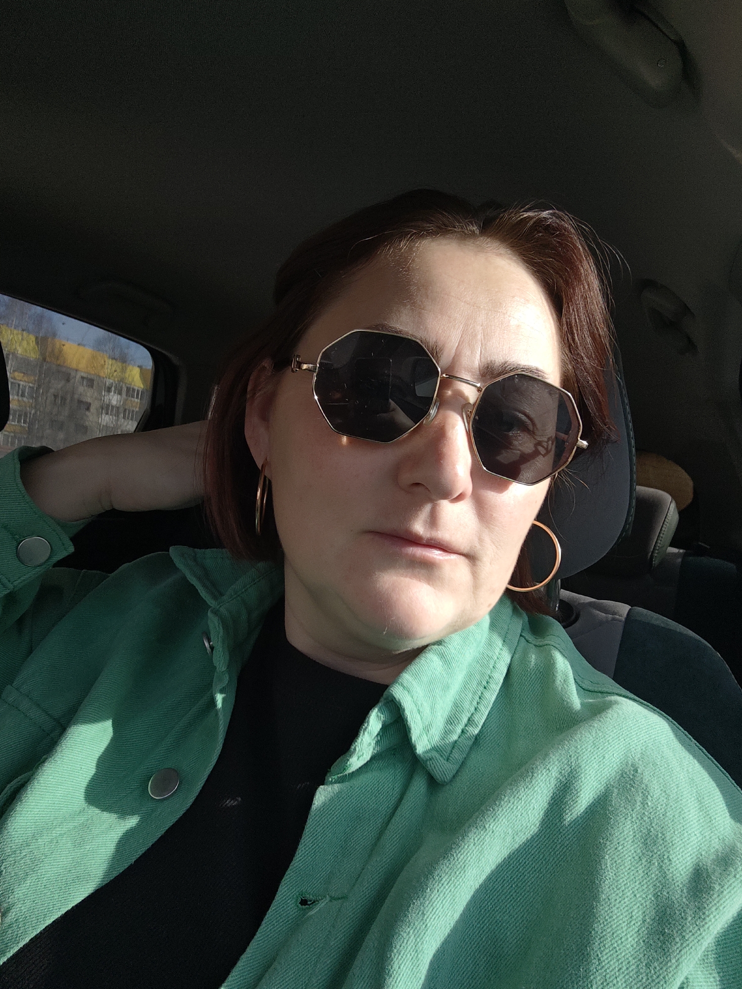 A female individual is depicted seated inside a vehicle, presumed to be in the city of Марковский, Russia. She possesses short, reddish-brown hair and wears dark, octagonal-framed sunglasses with gold-colored metal accents, paired with large, gold-colored hoop earrings. Her attire includes an open light-green collared jacket with silver-colored buttons worn over a dark-colored top. Her right arm is bent, with the hand resting behind her head. Direct sunlight illuminates her face and jacket from the front-right. Through the vehicle's left-side window, multi-story urban buildings, one featuring a yellow upper facade, and bare trees are visible in the background. The vehicle's interior headrest and dark upholstery are partially visible behind her.