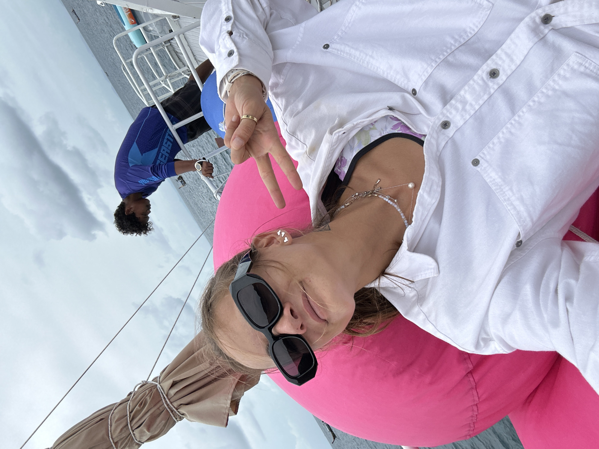 In Ao Nang, Thailand, on a boat with a white railing and a tan sail, a woman with blonde hair pulled back and large black sunglasses is sitting on a bright pink inflatable cushion. She is wearing a white button-down shirt over a patterned bikini top and a necklace with a pearl pendant. Her right hand is raised, with her index and middle fingers extended in a peace sign, and she is wearing a gold ring. Behind her, a man with dark, curly hair is leaning over the railing, wearing a blue long-sleeved shirt and dark shorts. The ocean is visible in the background under a cloudy sky.