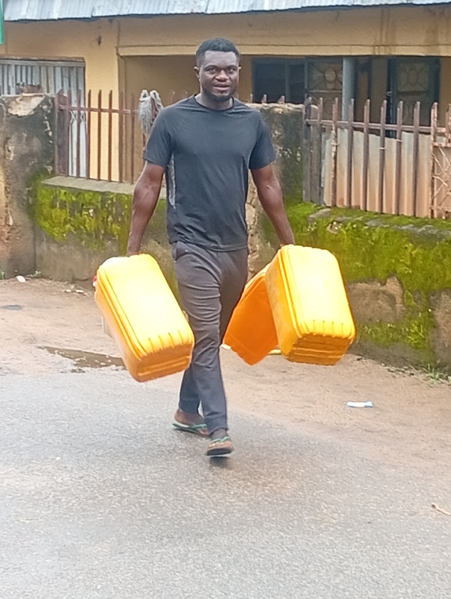 A Black man walks on a paved road in Jos, Nigeria, carrying two large yellow plastic containers filled with liquid. The man wears a black t-shirt and dark grey pants, with flip-flops on his feet. The road is wet, suggesting recent rain. Behind him, a weathered building with a metal roof is visible, with a rusty metal fence in front of it. Green moss grows on the wall of the building and along the fence. The scene appears to be in a residential area of the city.